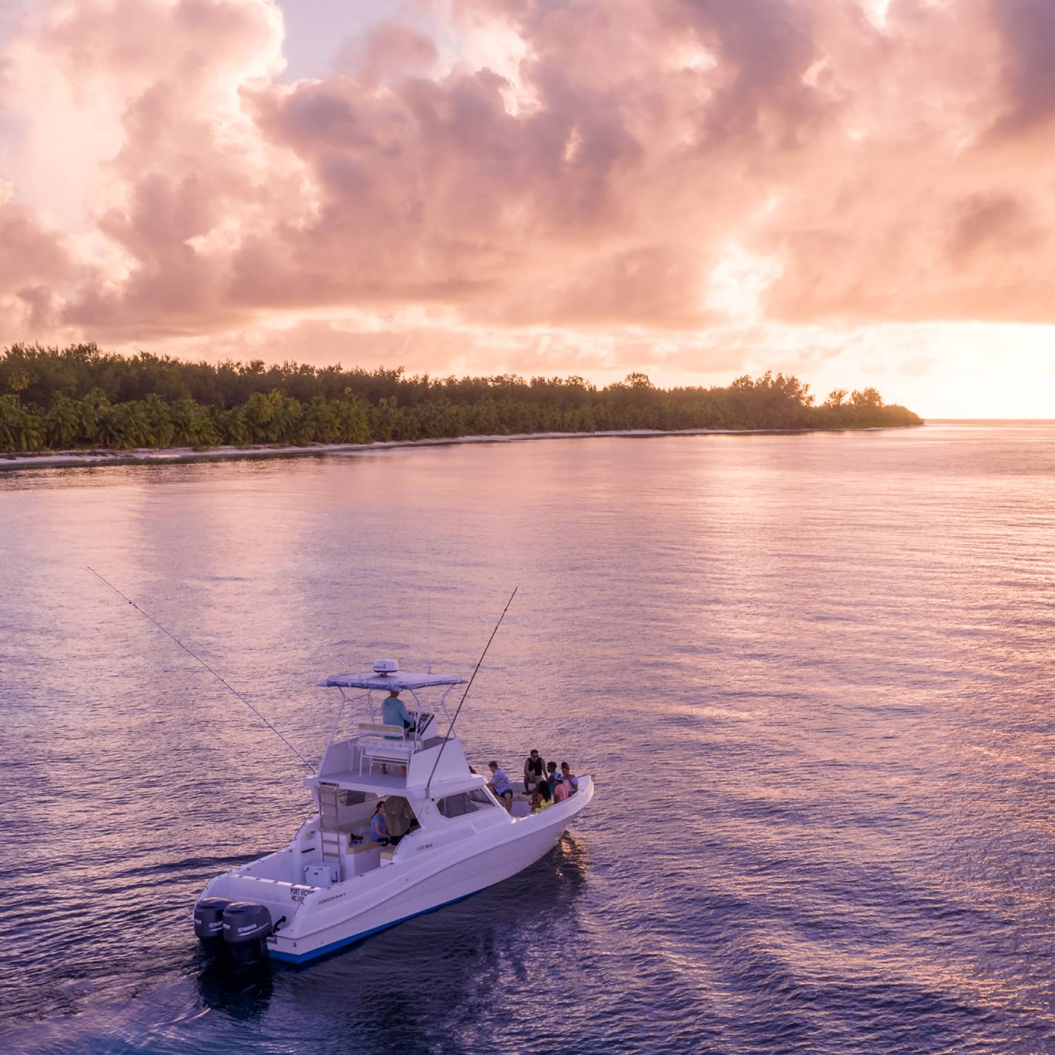 A group gathers at a boat's bow, drifting through calm water, a forested beach island and stunning sunset in the distance.