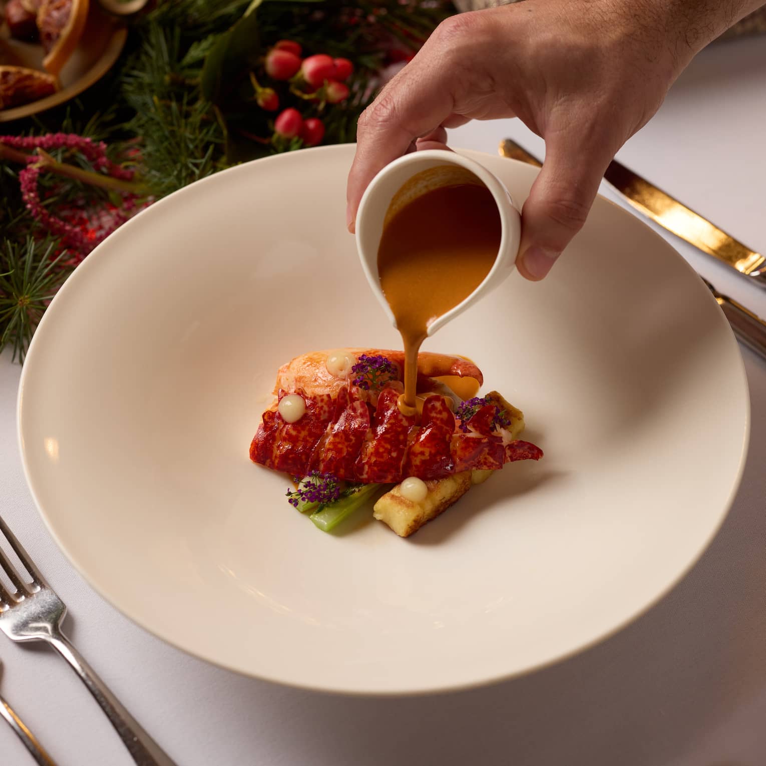 A person pours sauce over a food item in a shallow bowl set on a table decorated for the holidays.