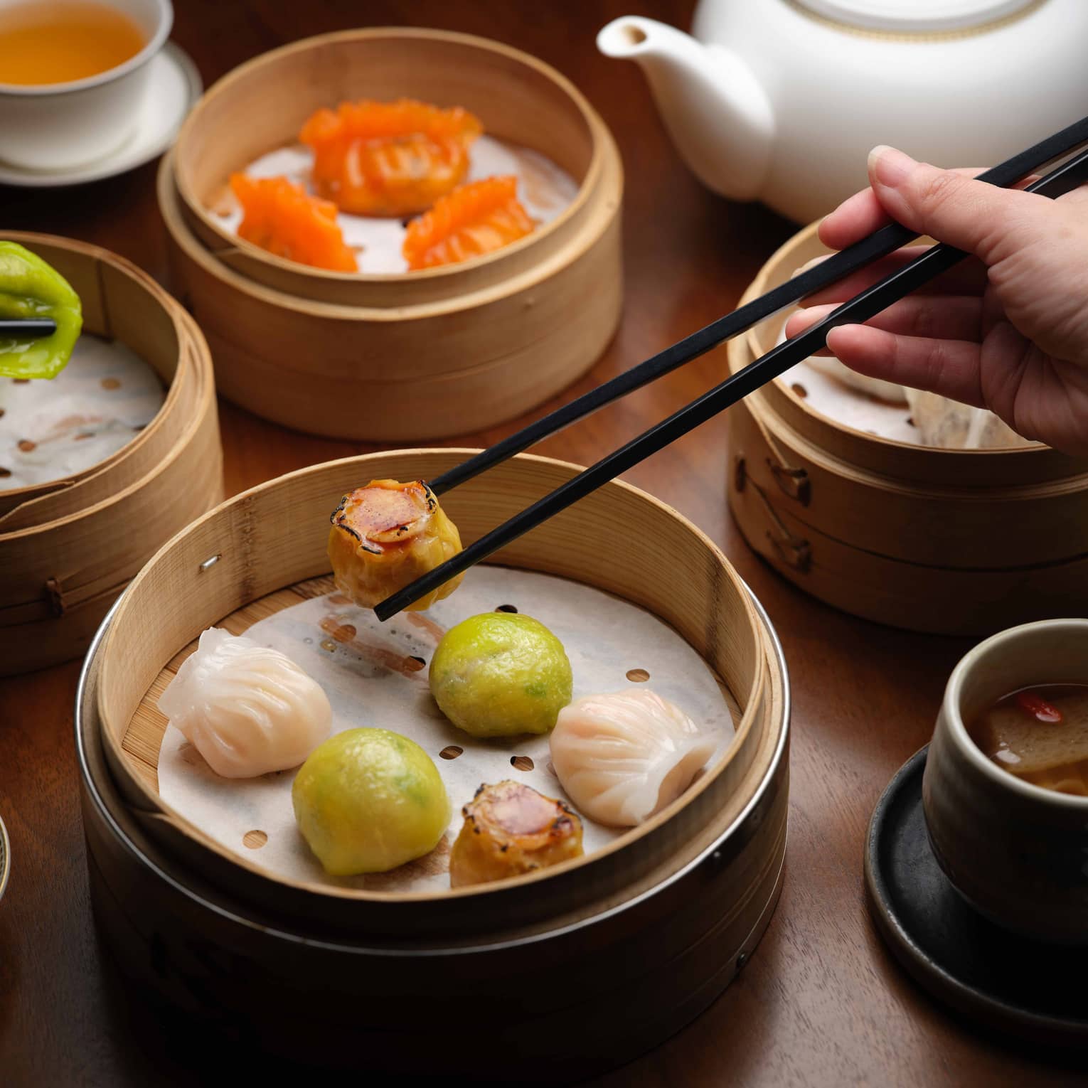 Beside cups of tea, a hand uses chopsticks to pick up one of the varied pieces of dim sum picked from nearby bamboo steamers.