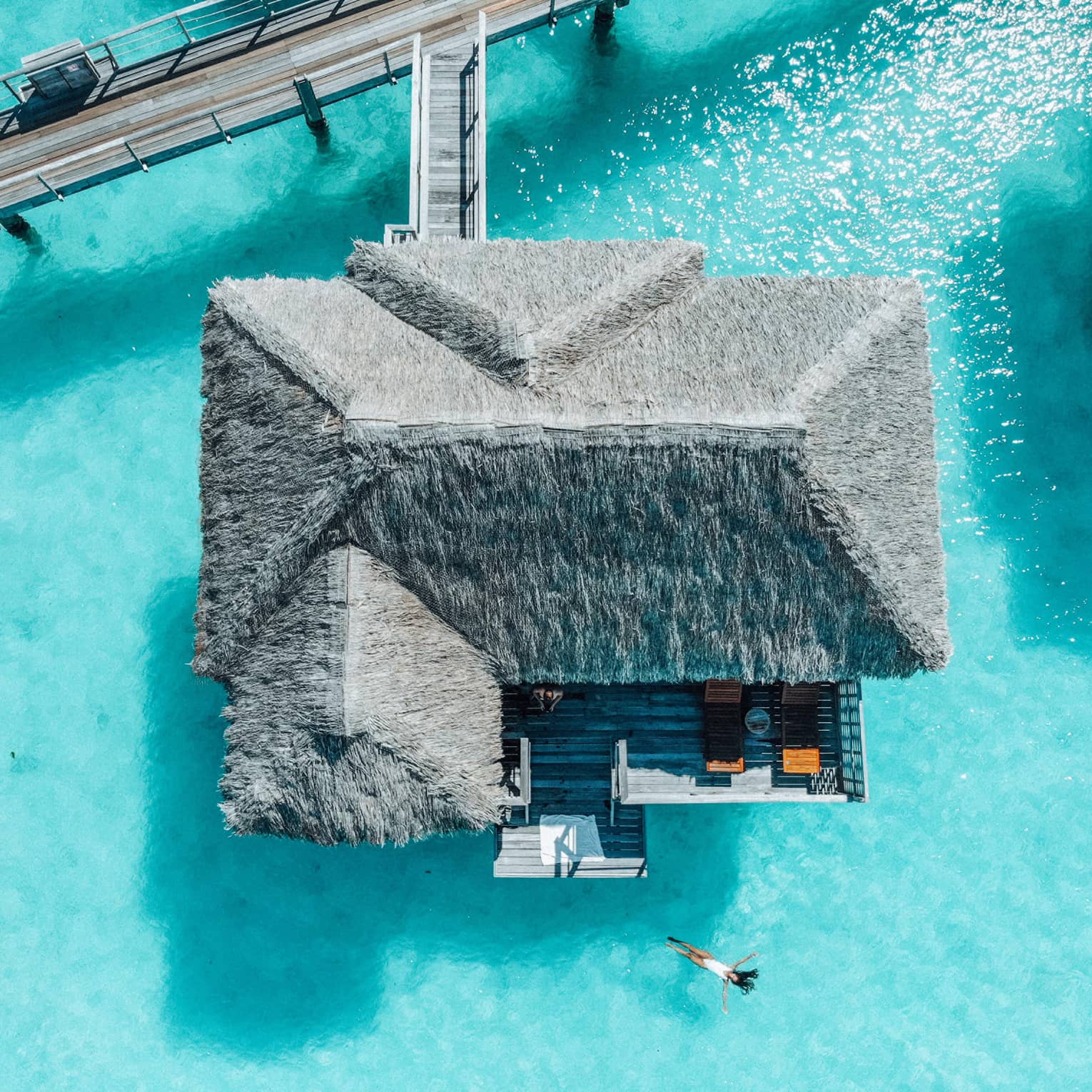 Aerial view of person swimming away from overwater bungalow in turquoise lagoon