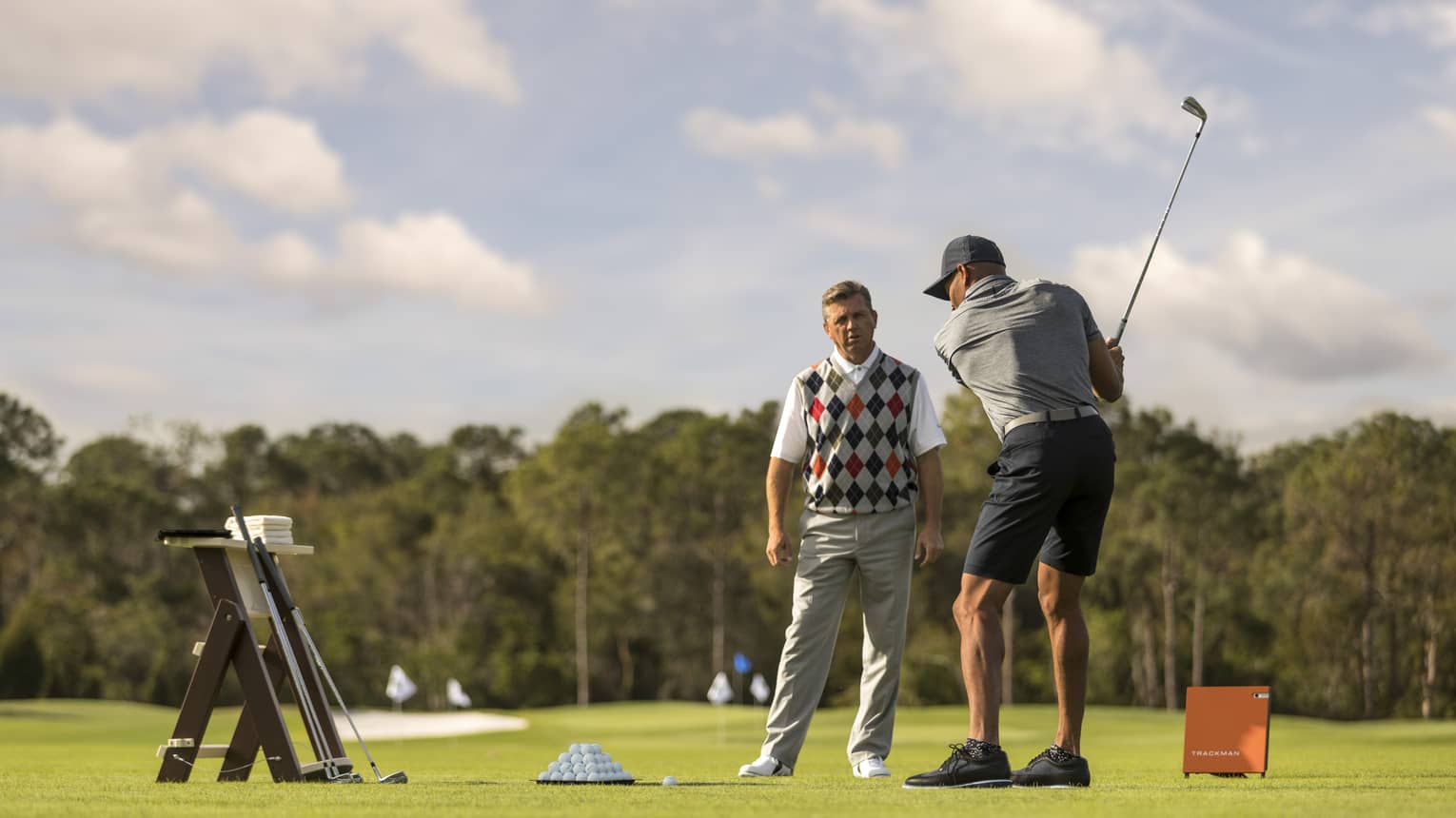 Against clear sky, one golfer looks on as another prepares to swing, a tray of balls and a stand of clubs nearby.