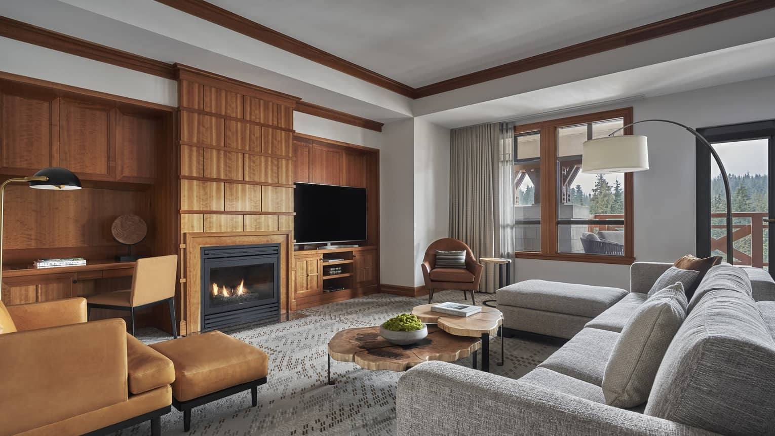 The Alpine Suite at Four Seasons Whistler featuring a sophisticated and cozy living area. The room is decorated with natural tones and includes a plush gray sectional sofa, a tan leather armchair with a matching ottoman, and wooden shelving units filled with books and decorative items. A contemporary fireplace and a large flat-screen TV mounted above it create a warm focal point. Large windows offer views of the mountainous landscape, contributing to the room’s tranquil ambiance.,The Alpine Suite at Four Seasons Whistler featuring a sophisticated and cozy living area. The room is decorated with natural tones and includes a plush gray sectional sofa, a tan leather armchair with a matching ottoman, and wooden shelving units filled with books and decorative items. A contemporary fireplace and a large flat-screen TV mounted above it create a warm focal point. Large windows offer views of the mountainous landscape, contributing to the room’s tranquil ambiance.