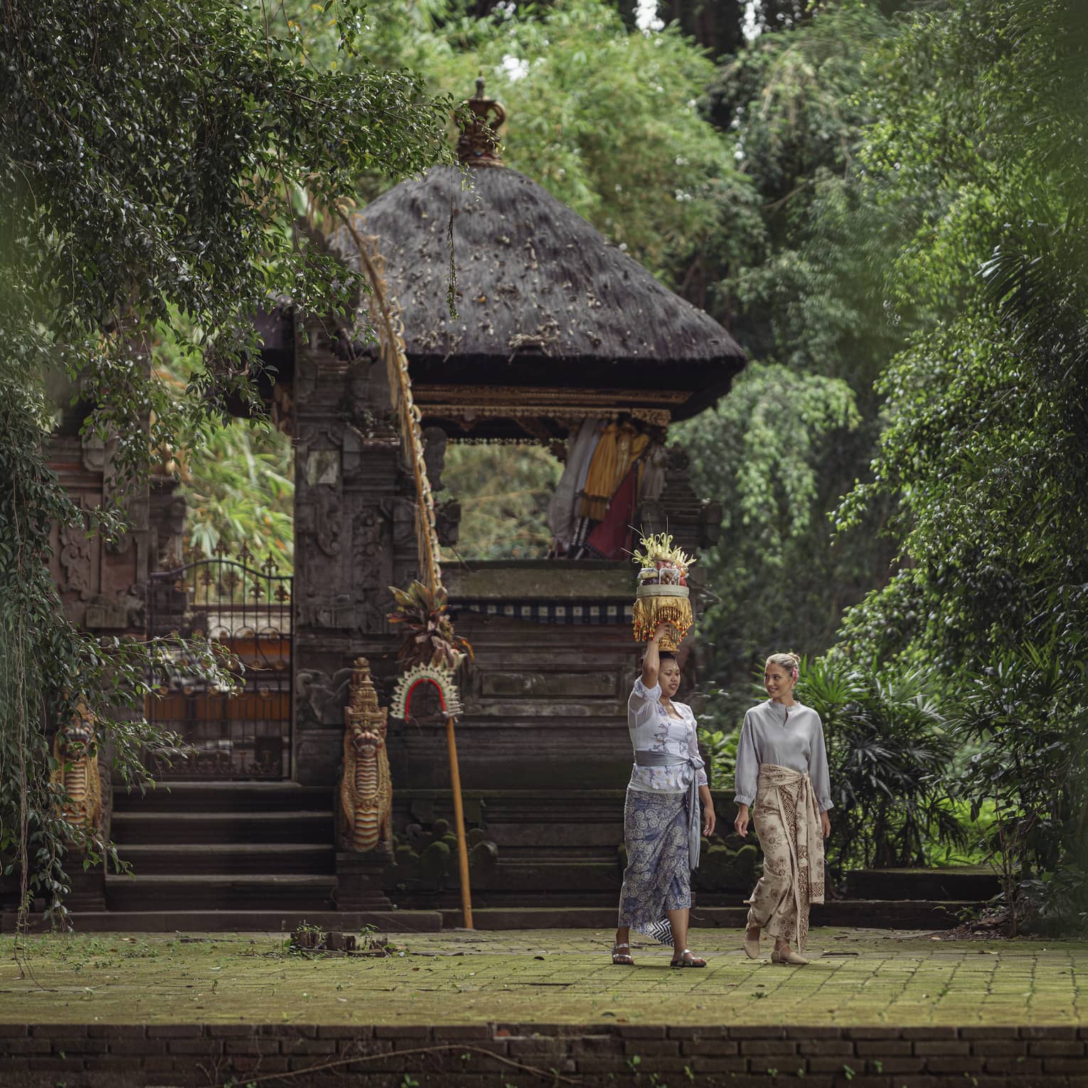 Two women wearing batik skirts walk by a temple in a lush tropical area as one of them carries a golden tray on her head.