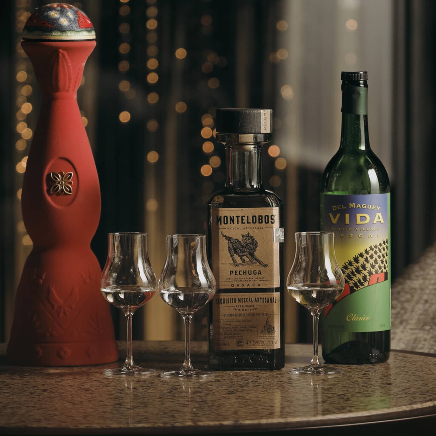 Tall, ornate red ceramic bottle alongside two more mezcal bottles and three tulip-shaped stemmed glasses of clear mezcal.