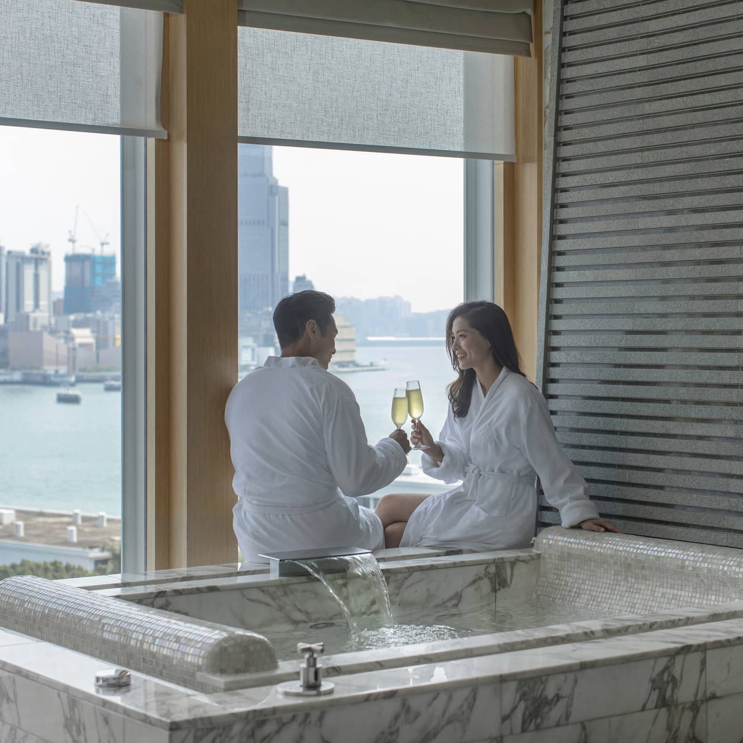 Man and woman clinking champagne glasses next to a private hot tub overlooking city harbour