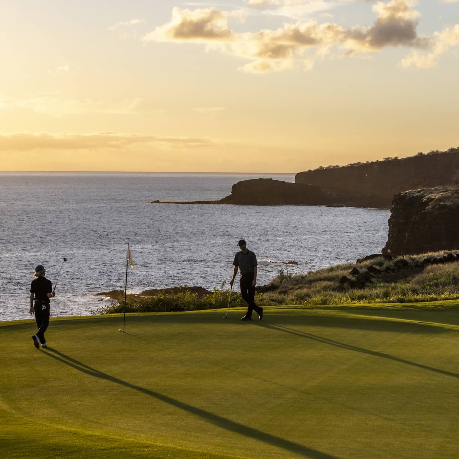 The setting sun silhouettes a pair of golfers enjoying a game of golf. The golf course is on top of a cliff beside the ocean.