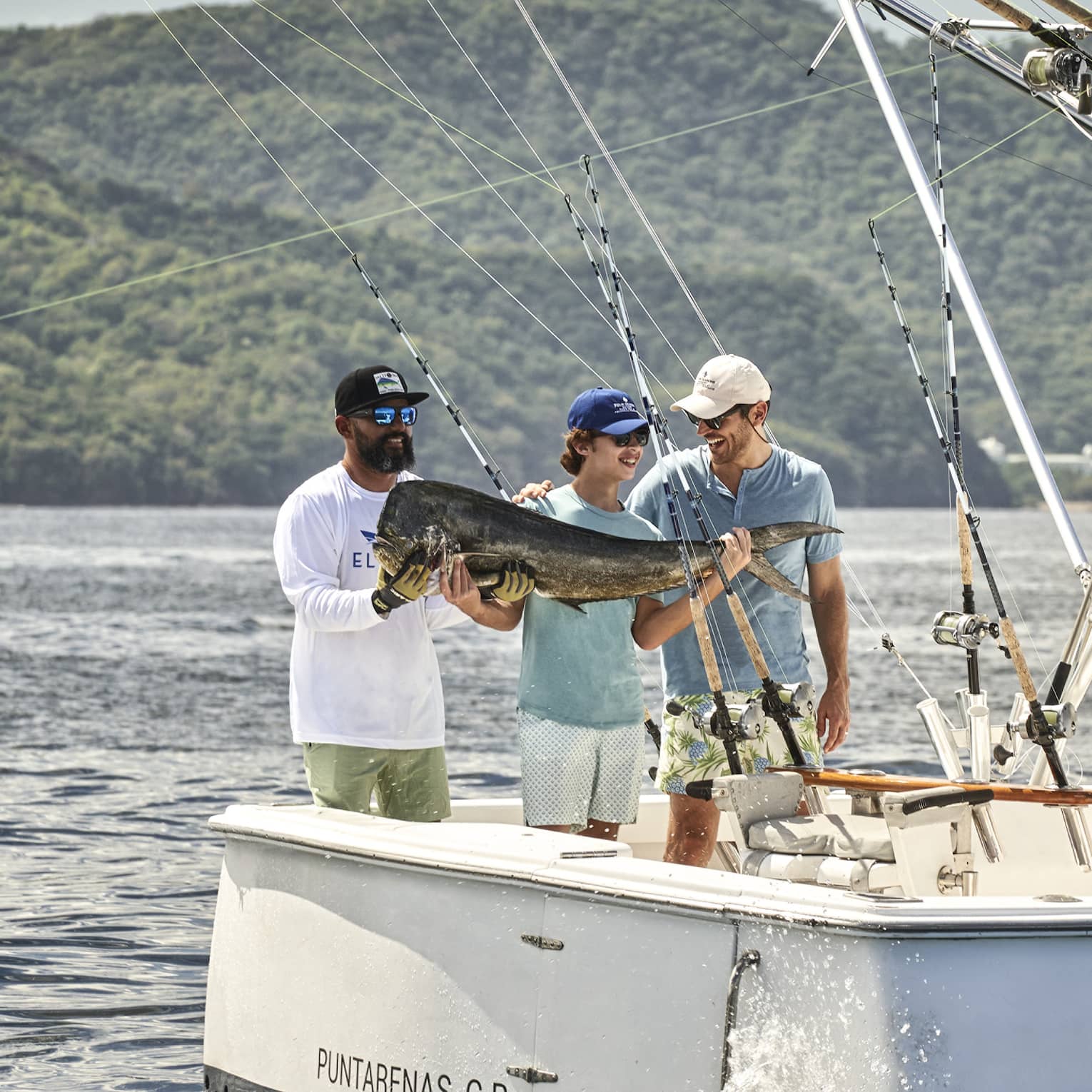Standing at the back of a fishing boat in the glistening sea against a lush mountain, three smiling people hold a huge fish.