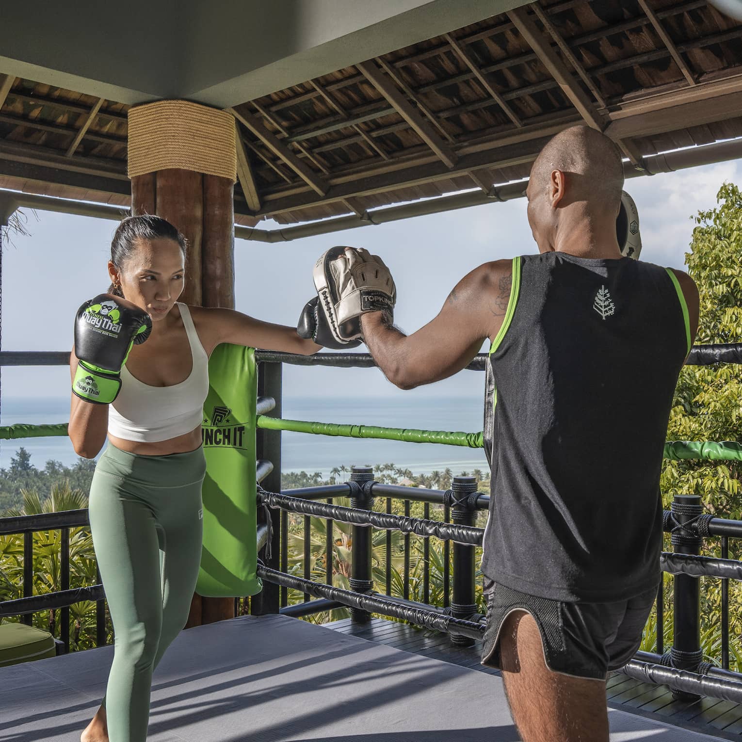 Two people practise Muay Thai in an outdoor boxing ring nestled in the treetops and overlooking the forest and the ocean.