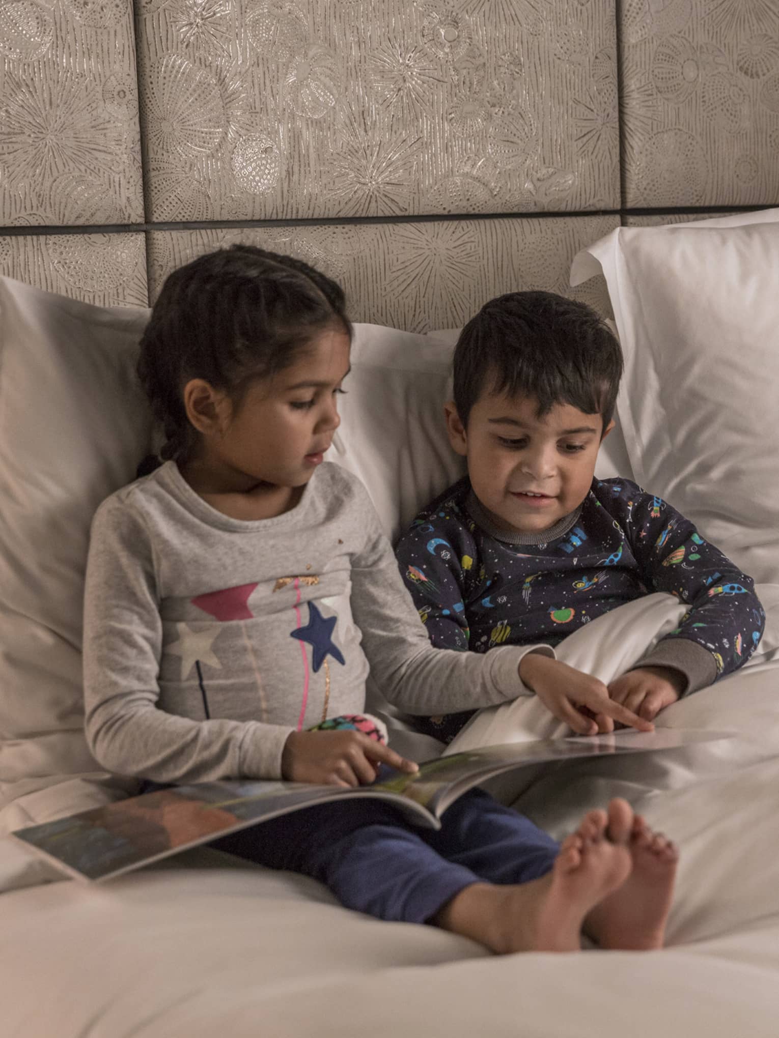 Young girl reads to her brother at bedtime, both in pajamas, a teddy bear propped on the pillow next to them