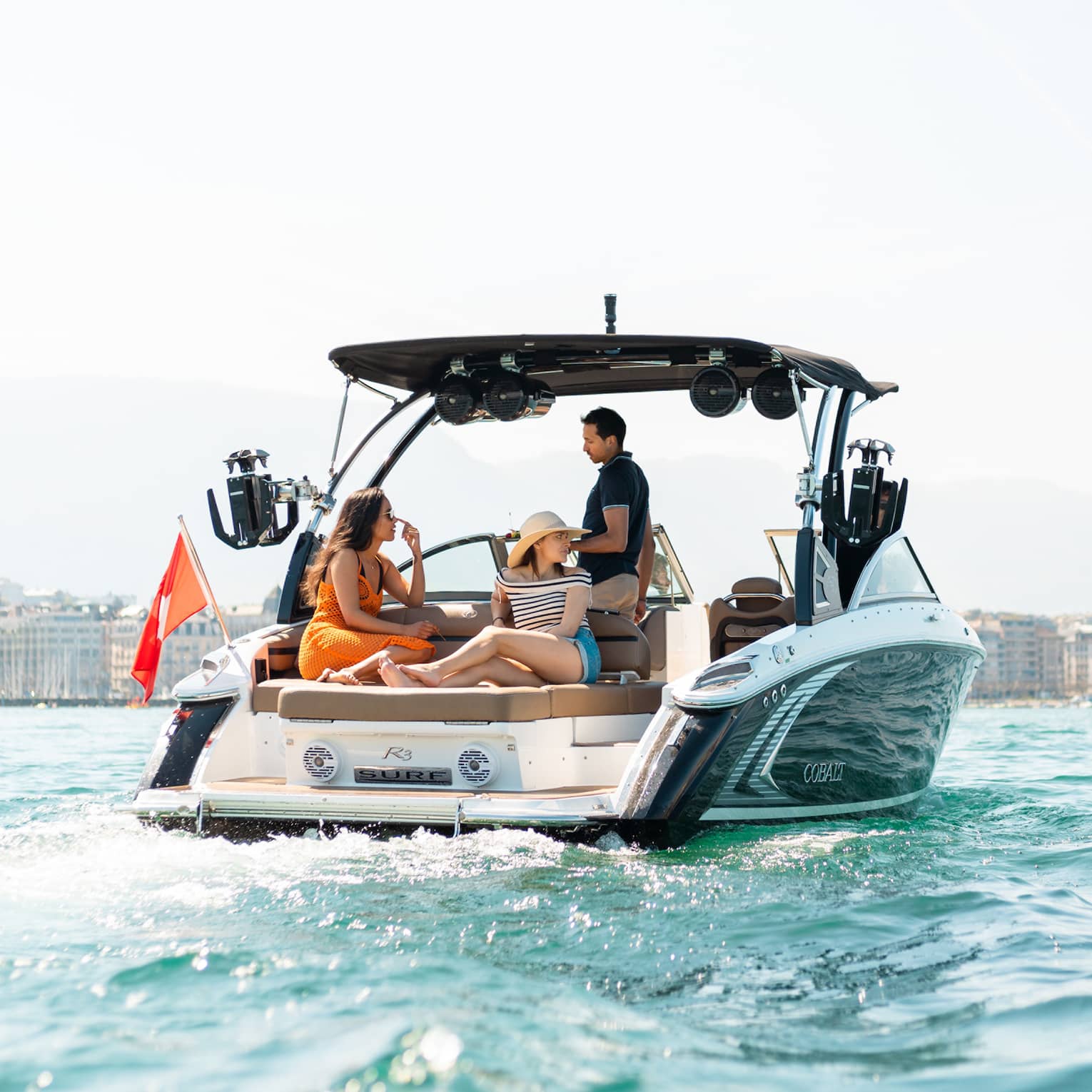 Captain guides a boat toward a building-lined shore while two guests lounge on a large cushioned seat behind him.