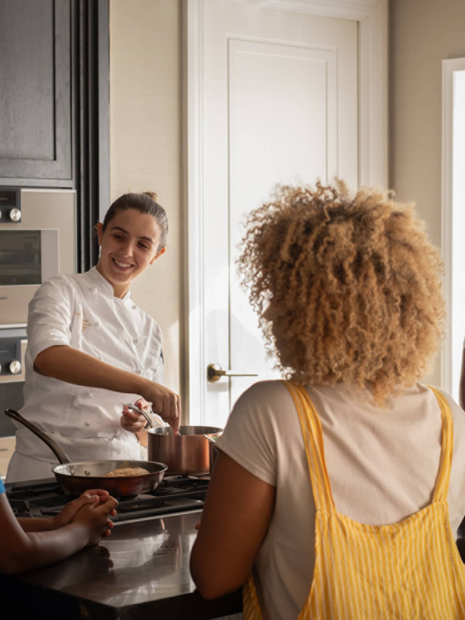 A family of four gathers around a kitchen-island cooktop watching a smiling chef prepare food in copper cookware.