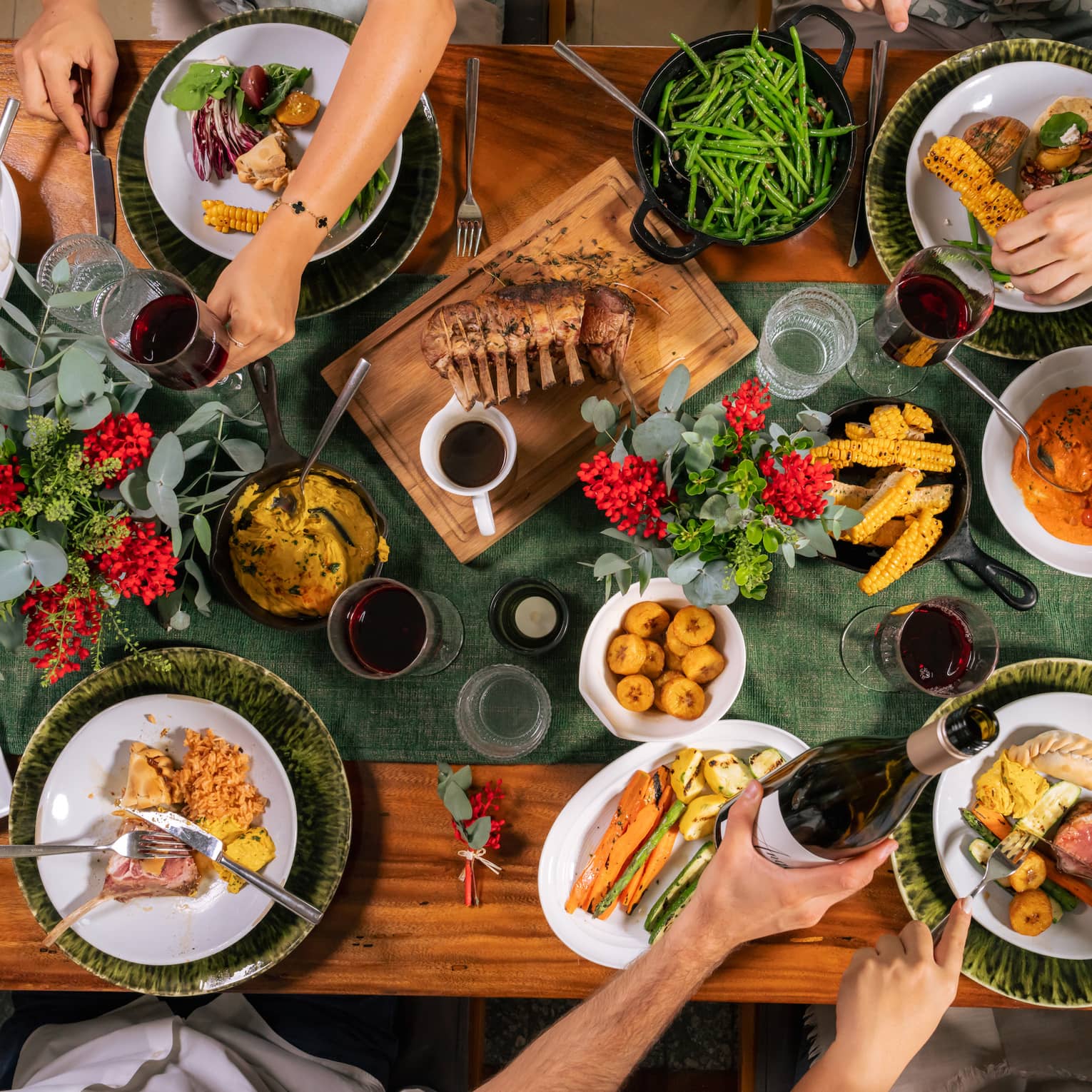Wooden rectangular dining table topped with dark-green runner, three vases of greenery and red berries and four plates of food as arms reach around to table to serve their plates