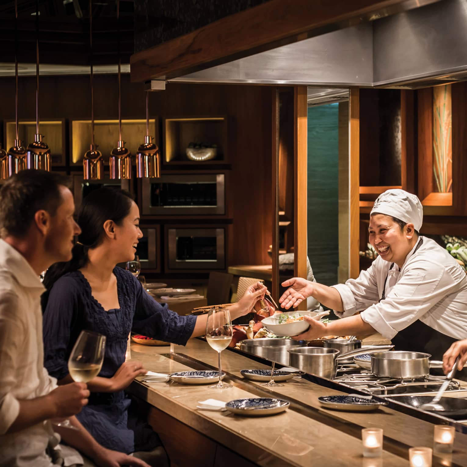Two chefs smile and laugh with a couple as they prepare them private dinner