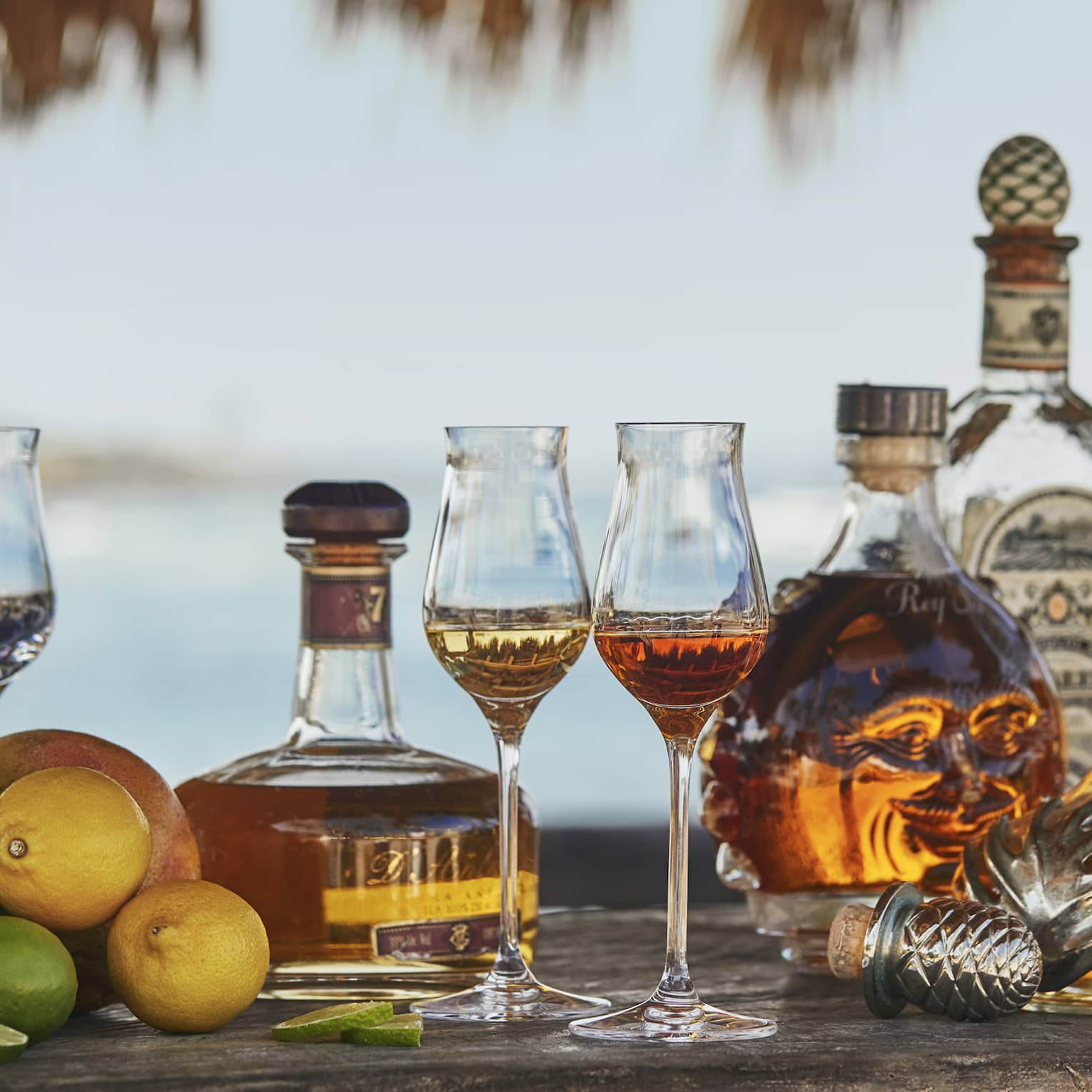 Whisky glasses surrounded by tequila, whisky and rum bottles, with a small pile of lemons and limes off to one side.