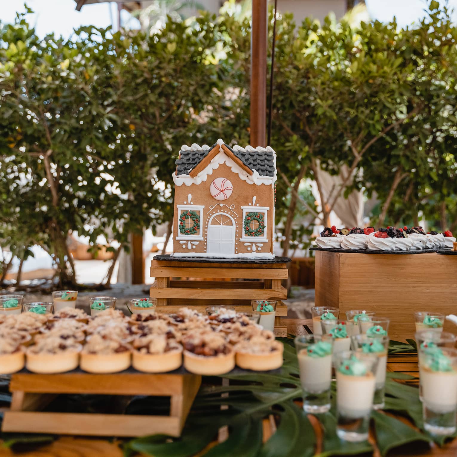 Wooden table set with holiday-themed desserts including cookies, tarts, merengues, custard-in-a-glass and a gingerbread house, all placed on pallet-style stands of varying heights