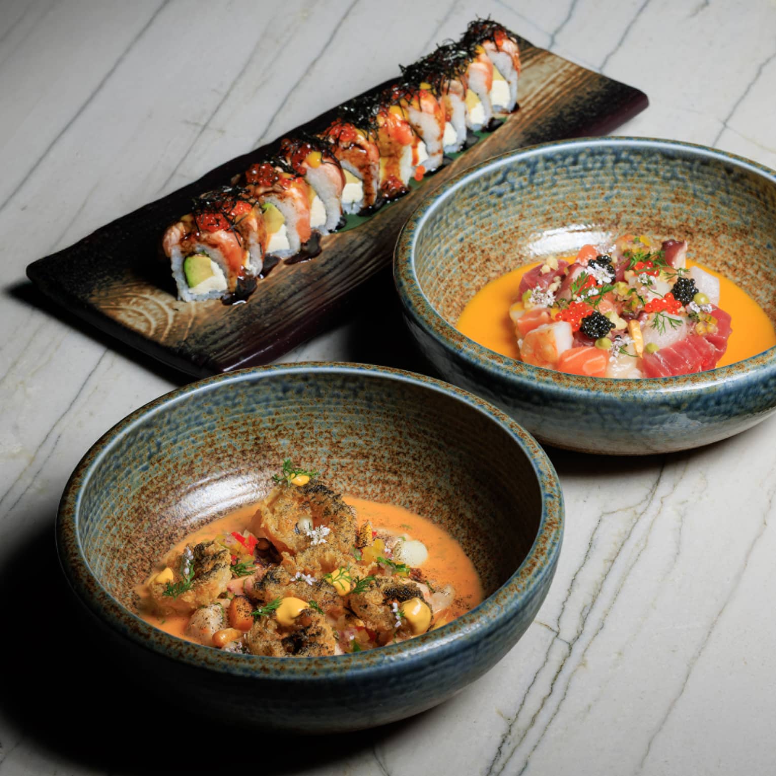 Two stoneware bowls filled with food sit next to a rectangular platter of sushi on a white wood-grain table