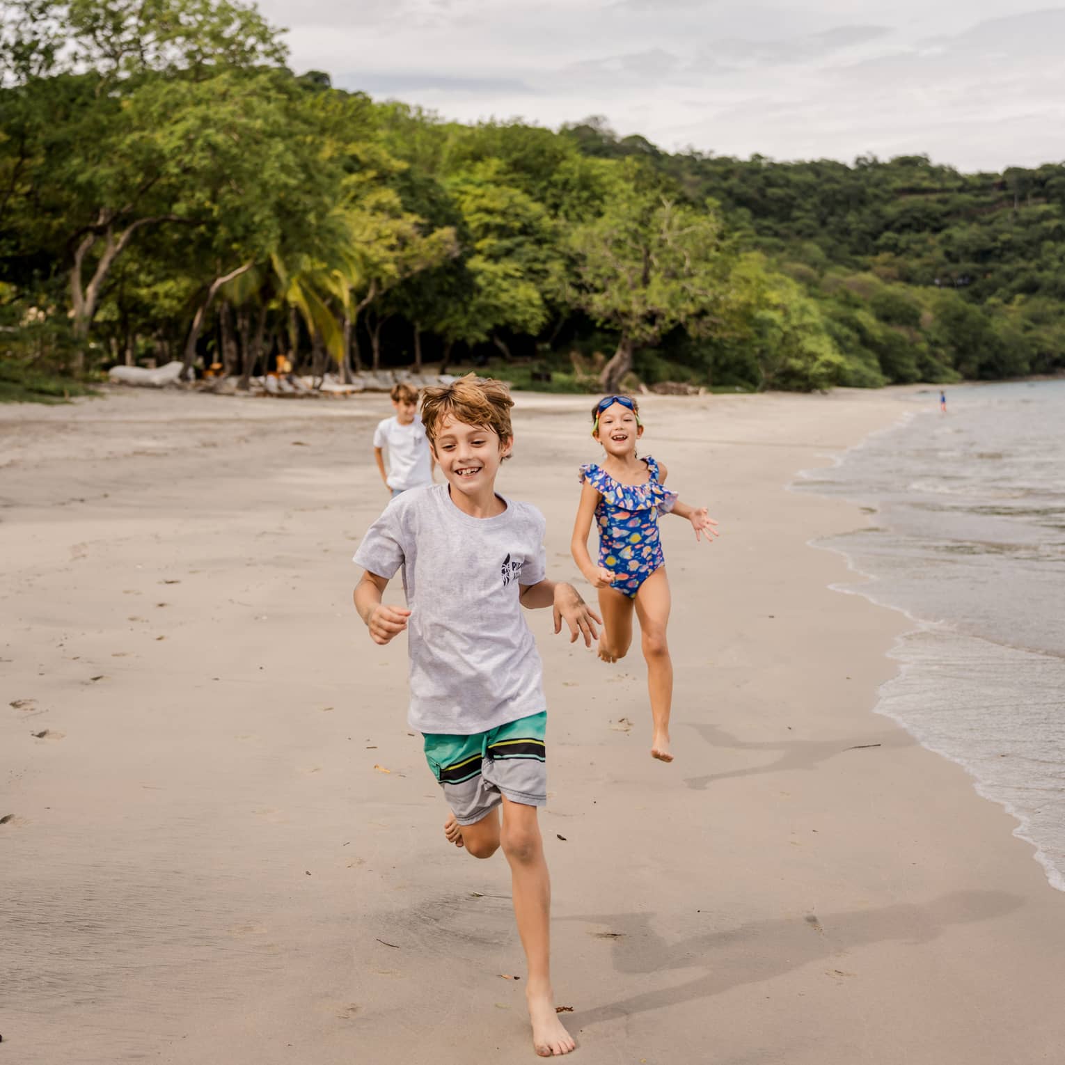Three children run along the beach with the ocean on one side and a lush treeline behind them