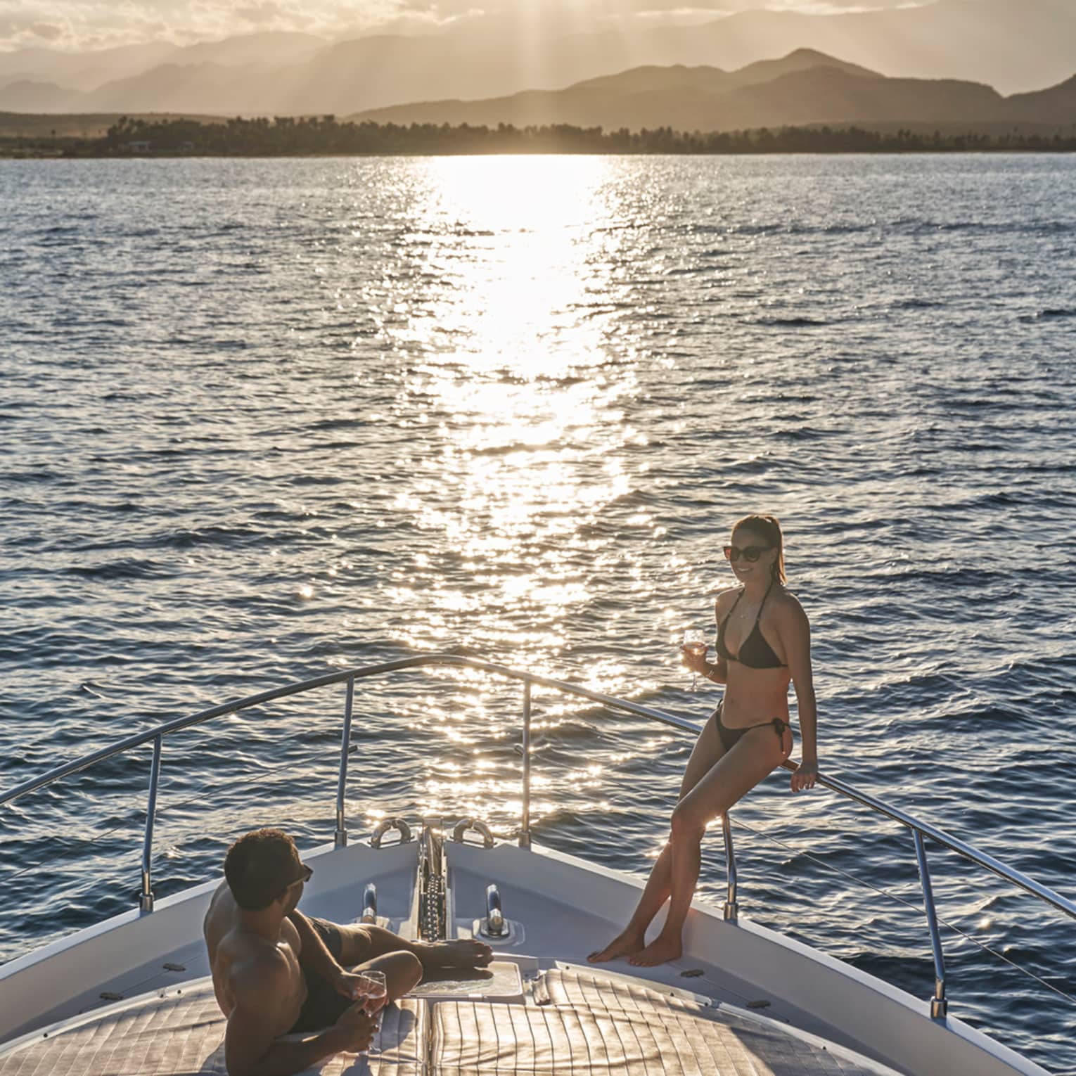 A couple in swimwear relaxing on the bow of a boat, wine glasses in hand and the setting sun glinting off the endless ocean.
