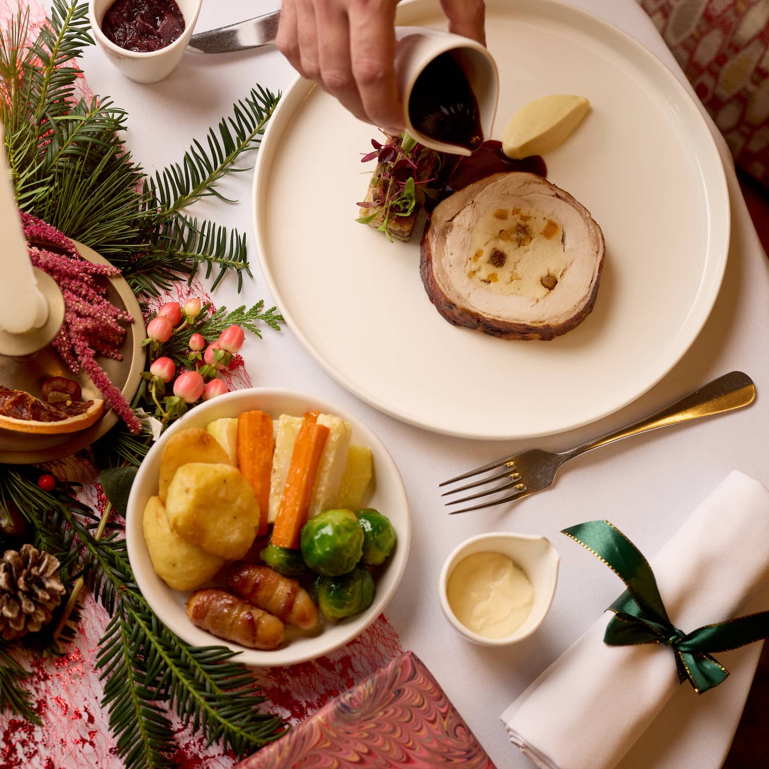 A table set with holiday dishes and a holiday centrepiece. A person pours sauce on a plate of food items.