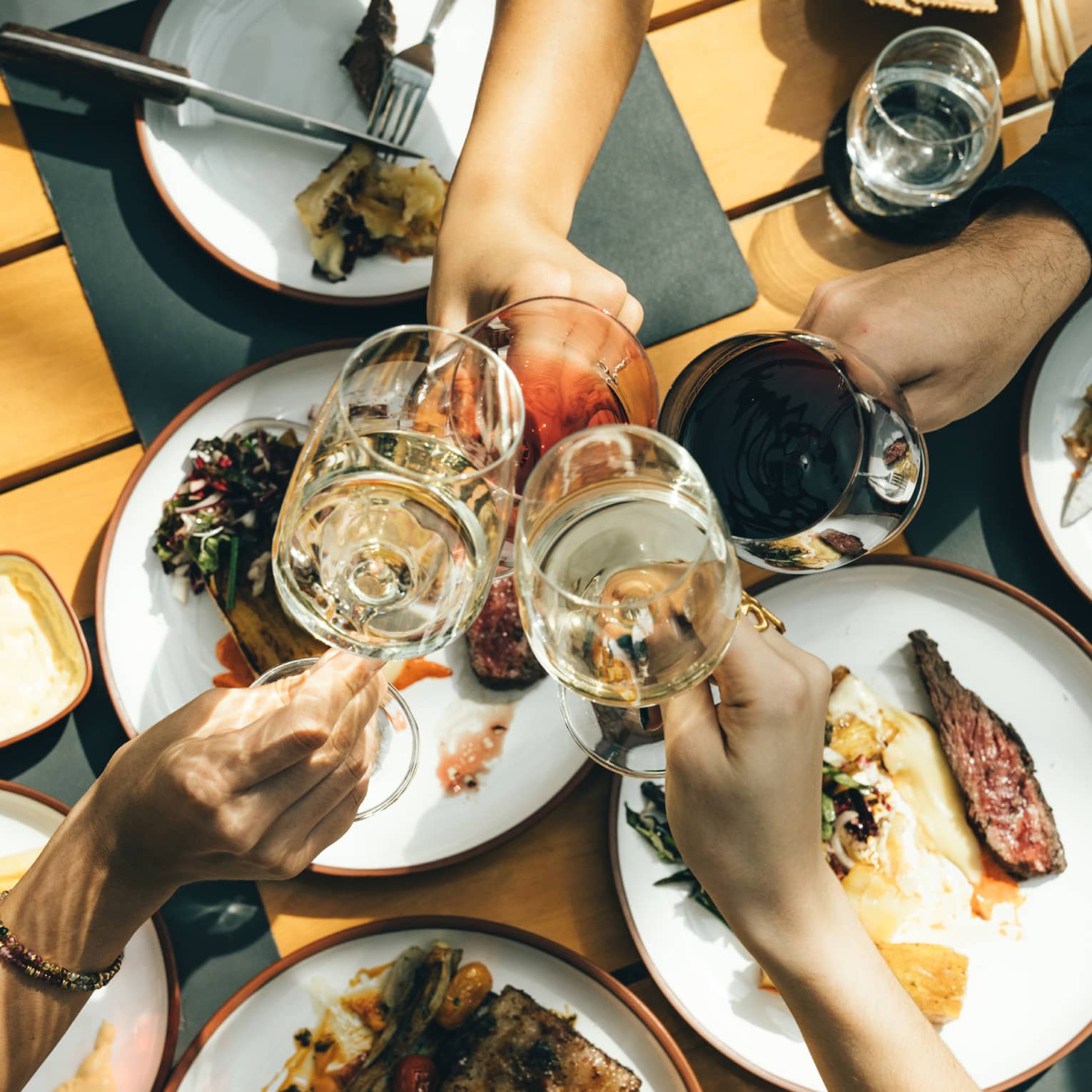 Close-up from above of four hands clinking glasses at the centre of a spread of plates of rare grilled meat and vegetables.