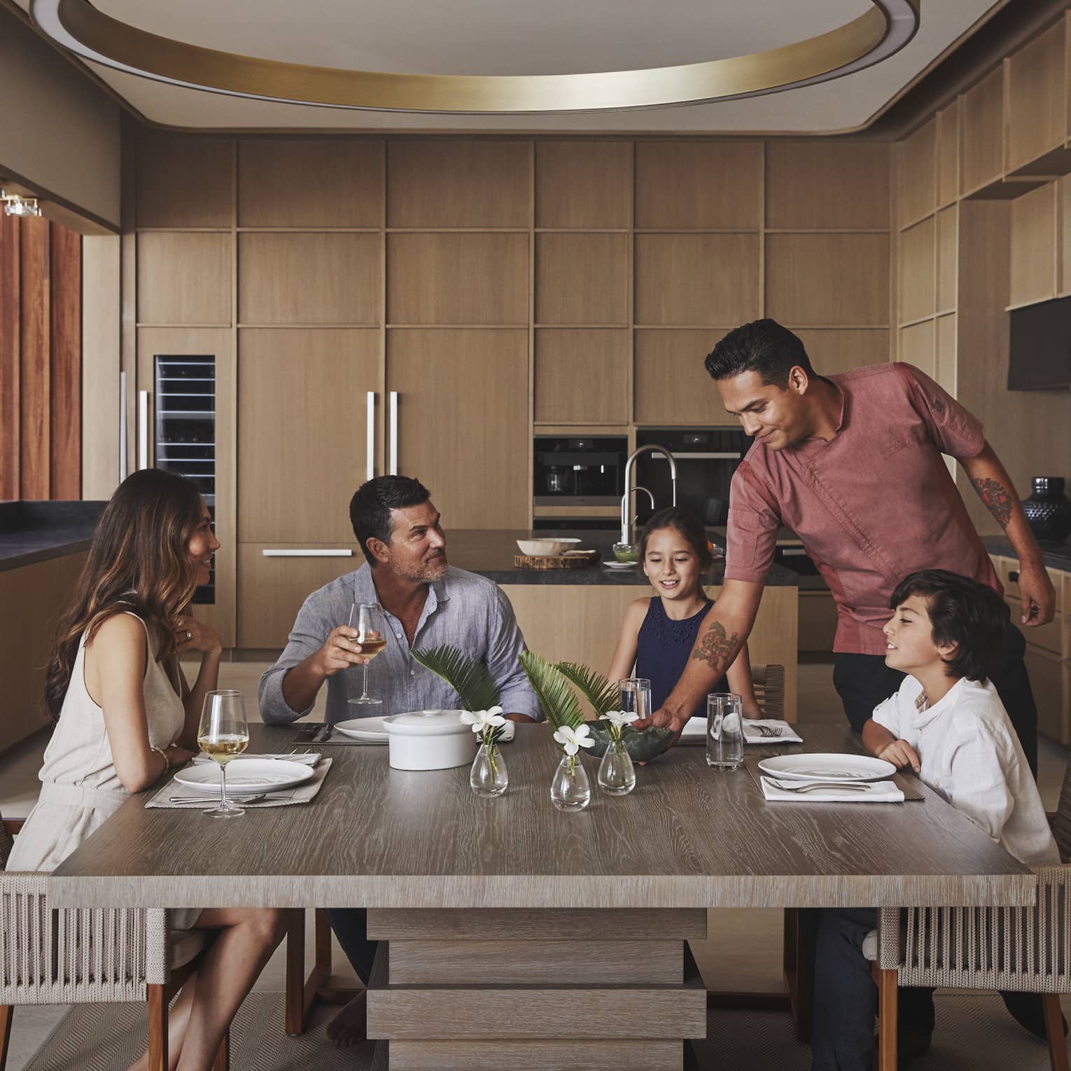 Four guests sitting at the table in a villa while a chef places a dish on the table.