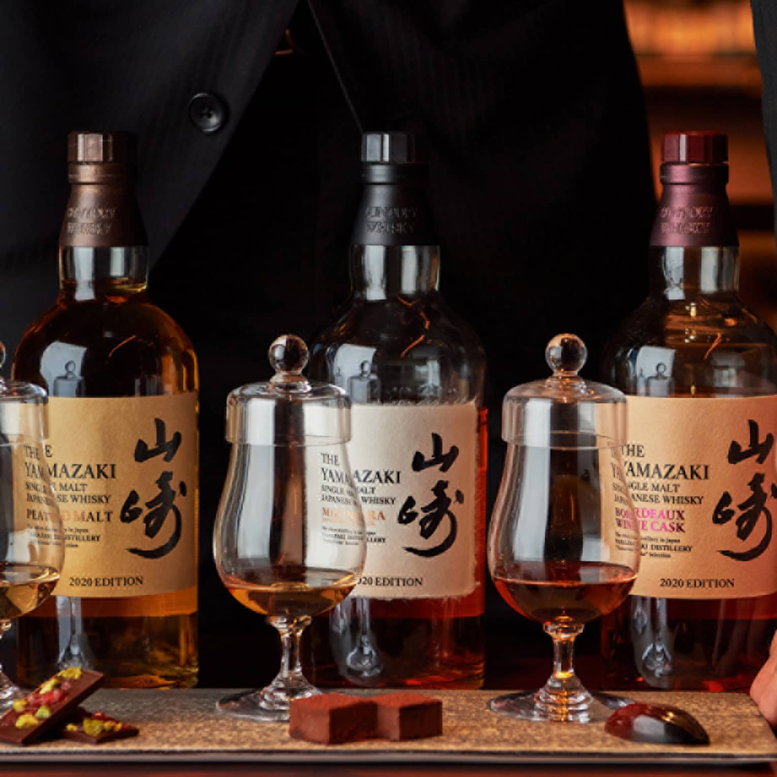 Hands frame a row of three bottles of single malt Japanese whisky behind three curved whisky tumblers with tasting caps.