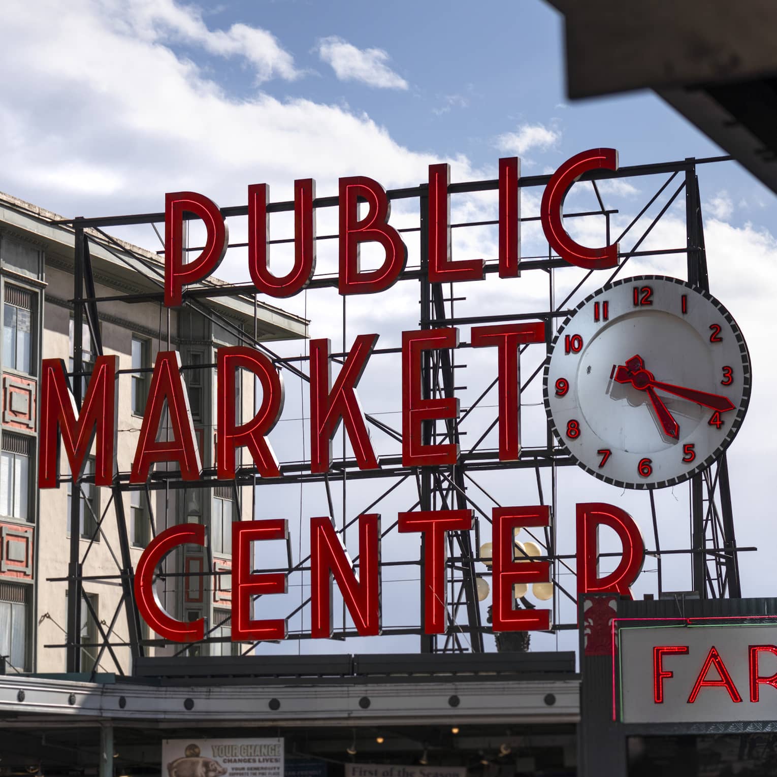 The giant Public Market Center clock and sign set in red neon letters on black scaffolding against a blue sky with clouds.