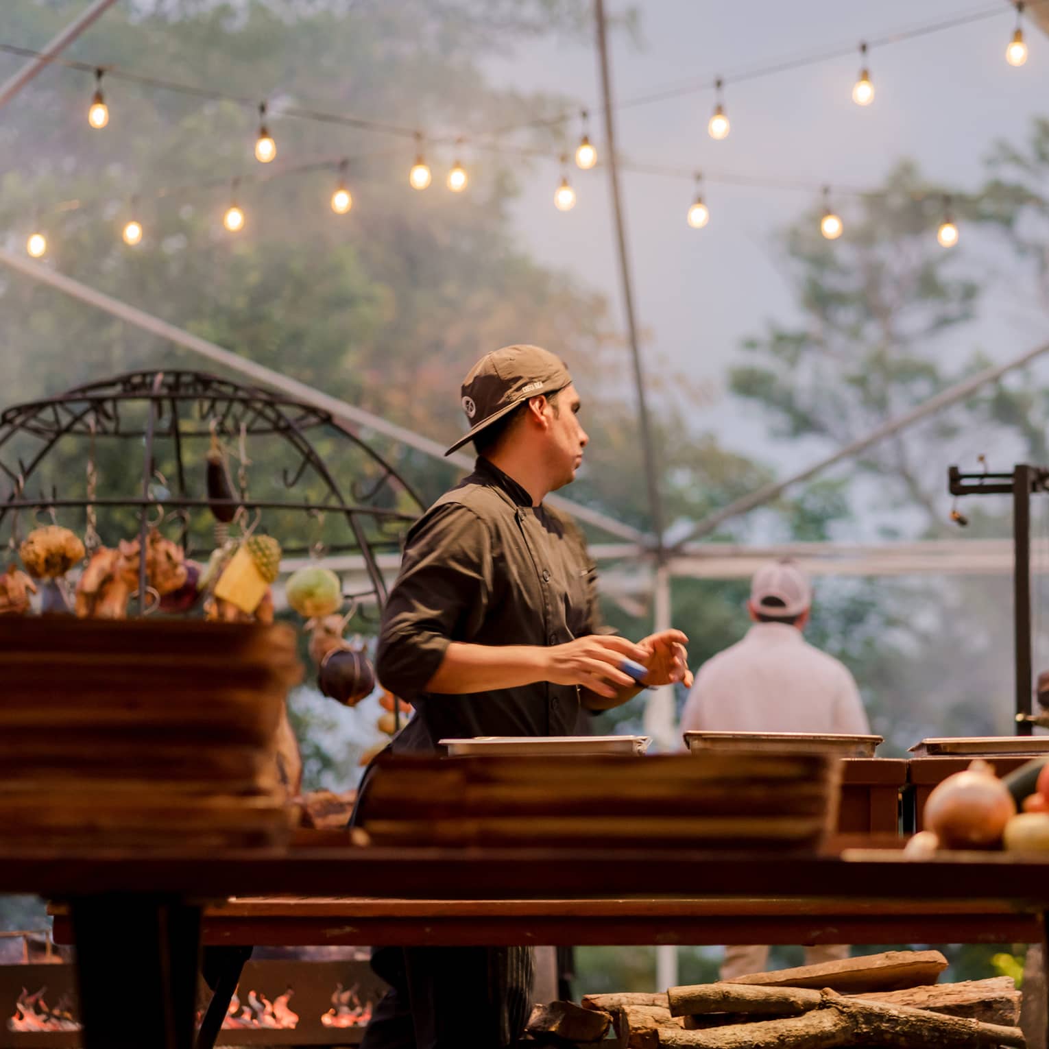 Chef wearing a black chef's jacket stands a grilling station beneath two strings of twinkling lights