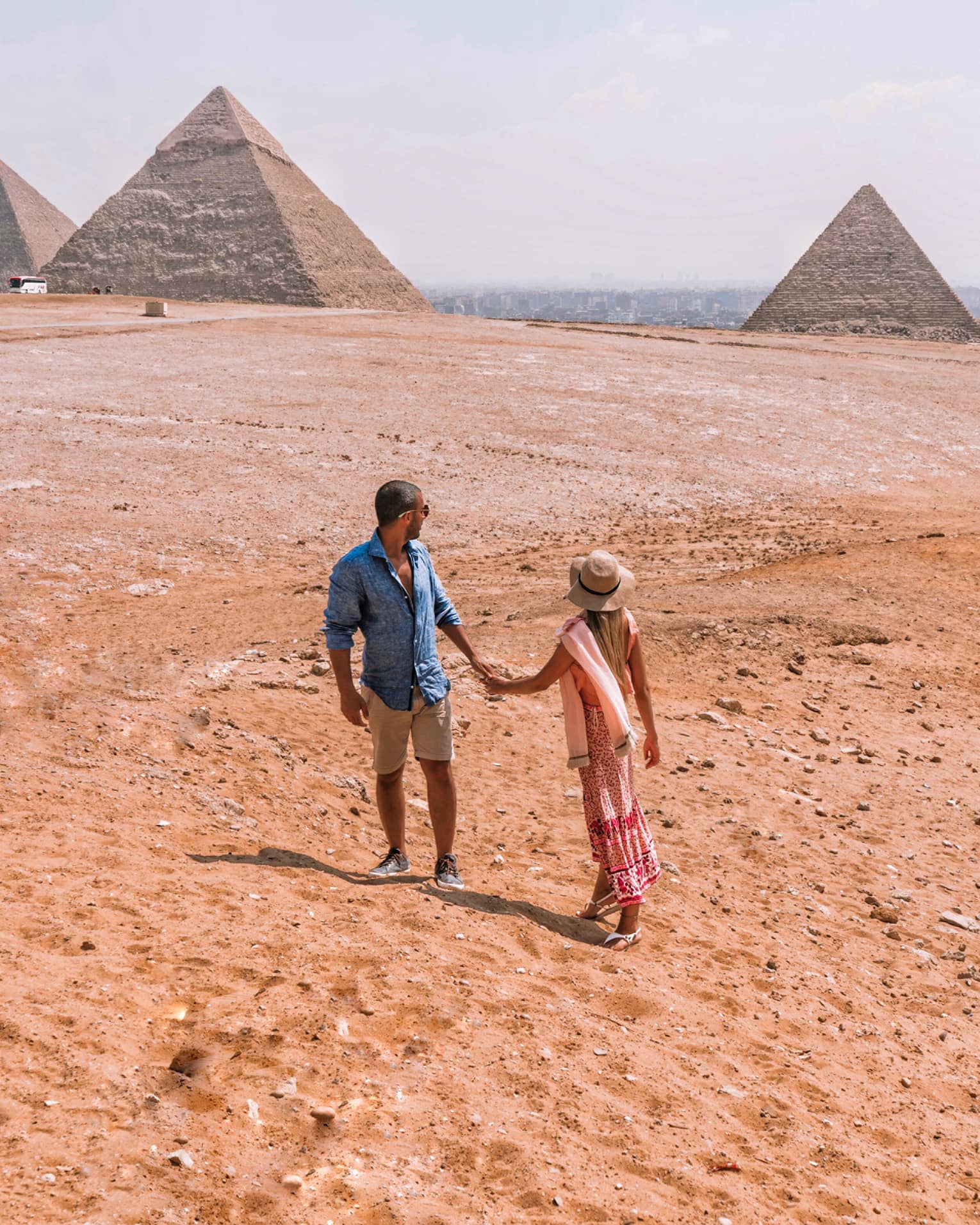 A couple holding hands turn back to look across the arid desert at the giant pyramids in the distance.