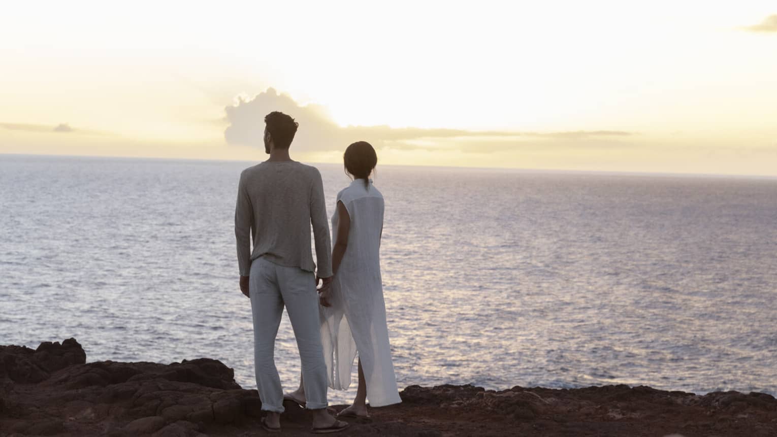 Rear view of a couple standing on a rocky ledge overlooking the vast ocean as they watch the sun set behind distant clouds.