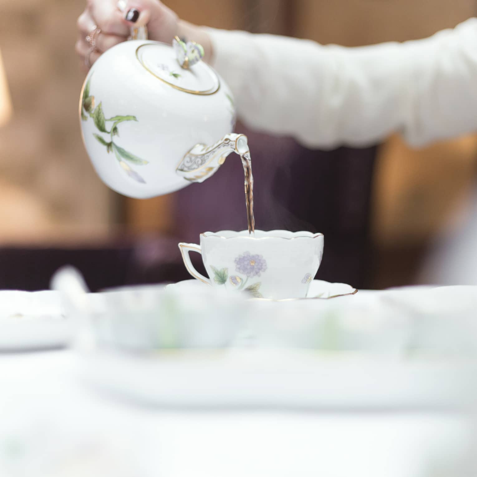 Person holds floral tea pot and pours tea into cup and saucer