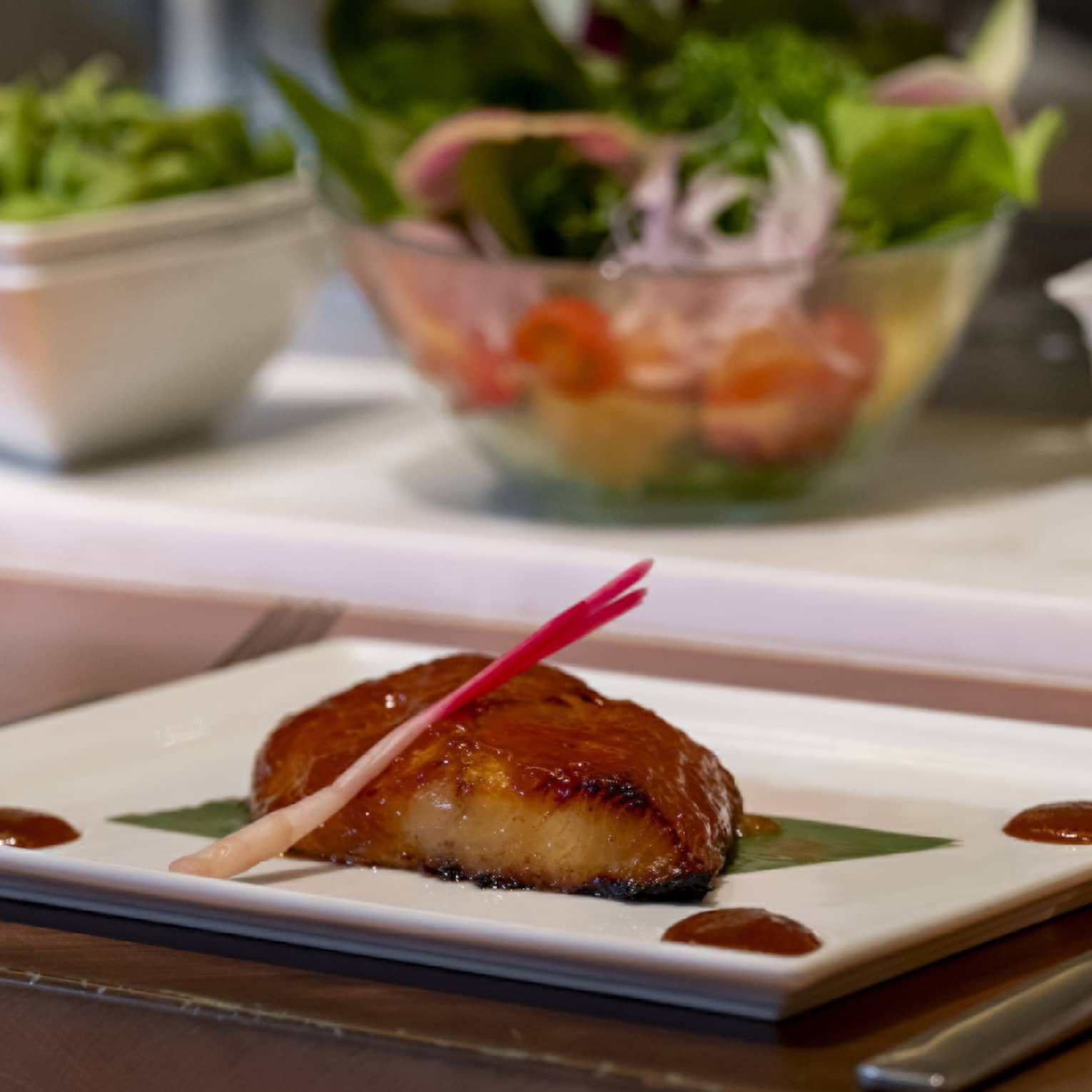 Roasted miso black cod centered on a rectangular plate and pickled ginger sprout garnish, dots of brown sauce in each corner.