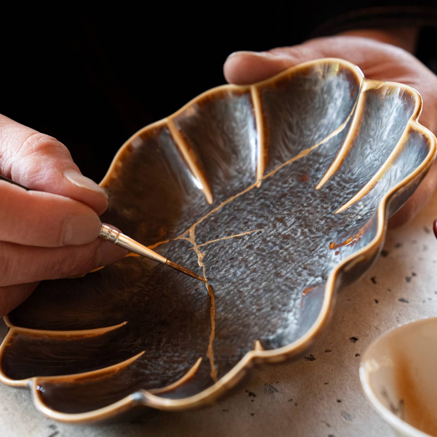 A person using the art of Kintsugi, by using gold paint to paint over the cracks of a once broken black flower shaped plate.