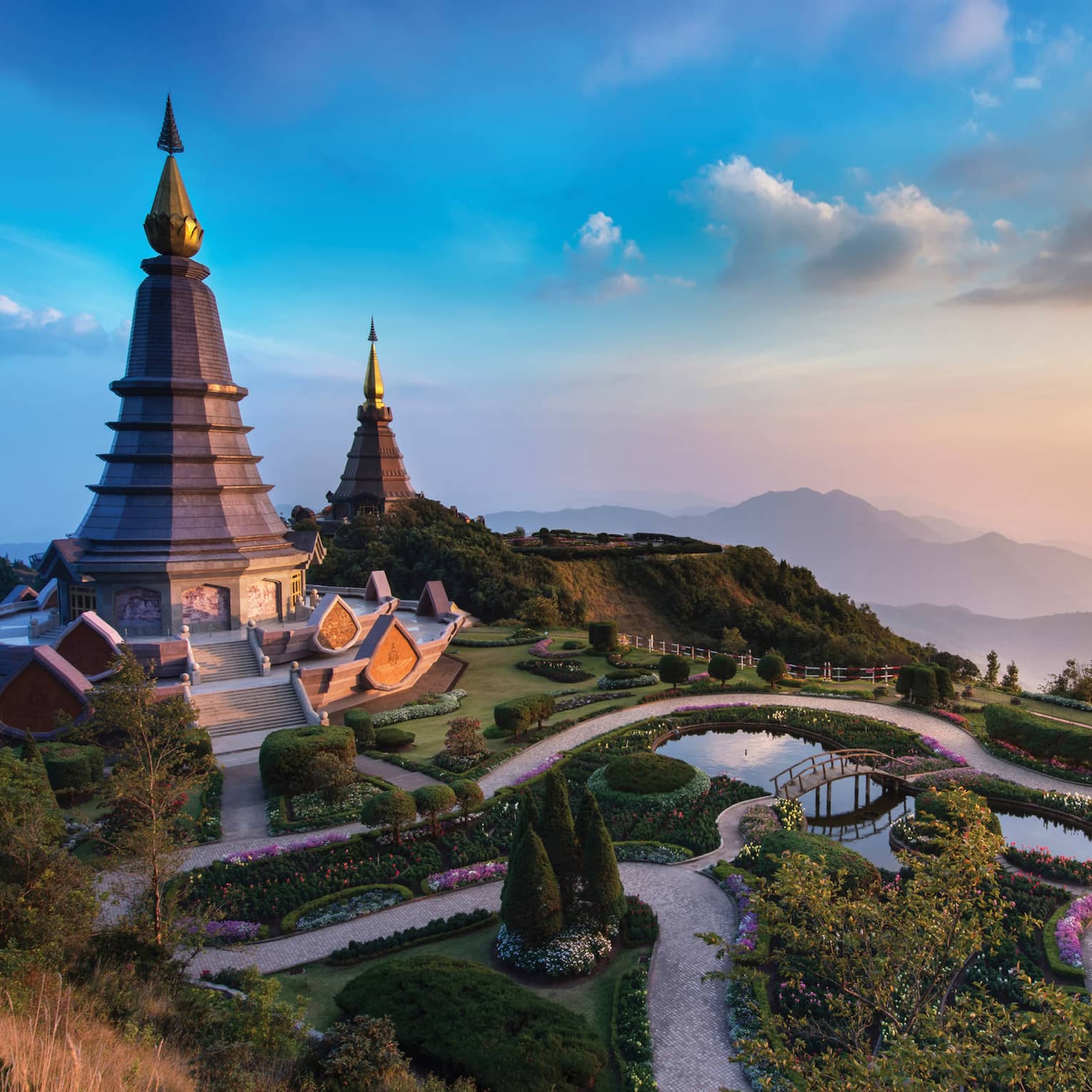 Majestic twin pagodas surrounded by lush gardens and winding paths, set against a backdrop of misty mountains at sunset.