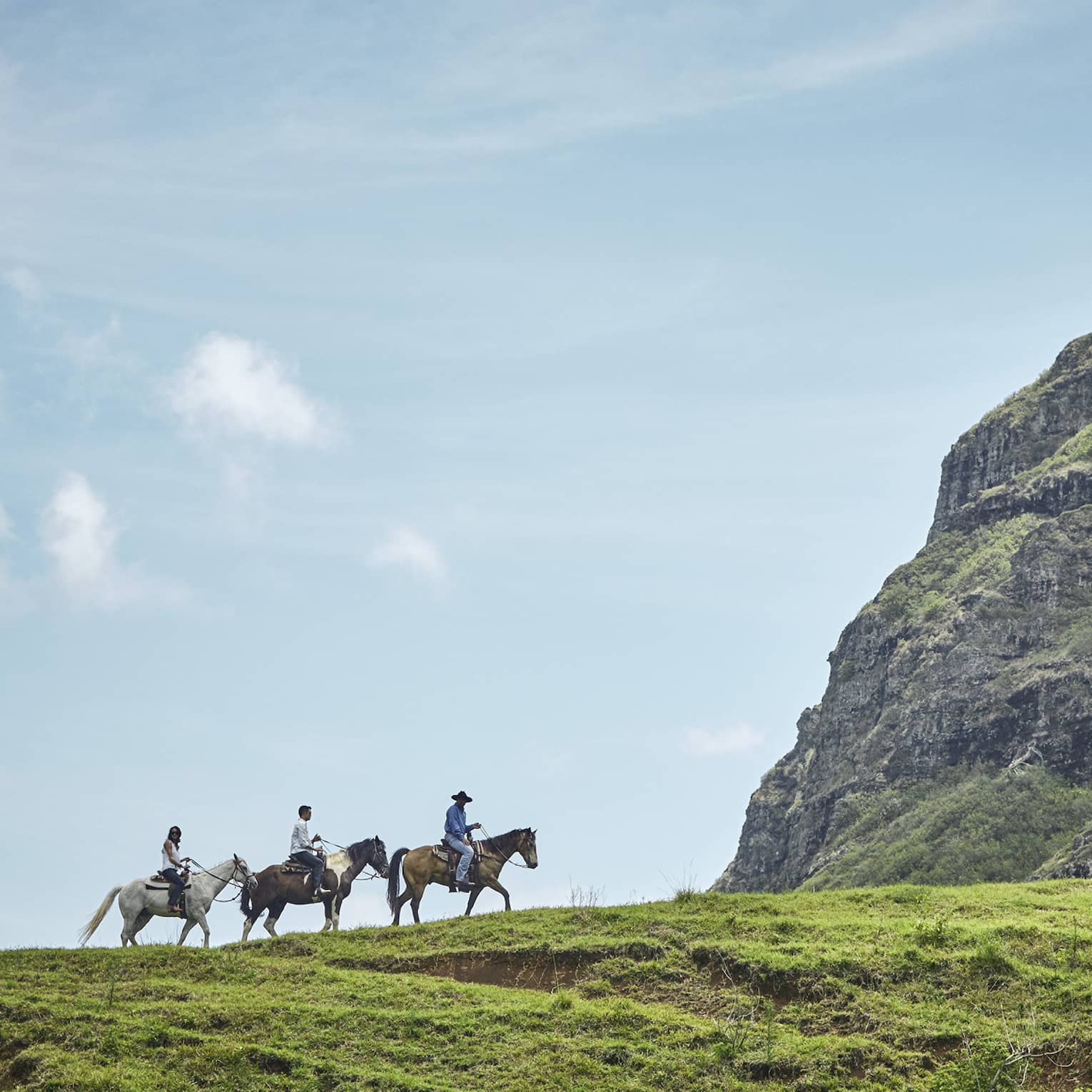 Long side view of three horseback riders on a grassy hill heading toward an imposing verdant cliff under clear blue skies.