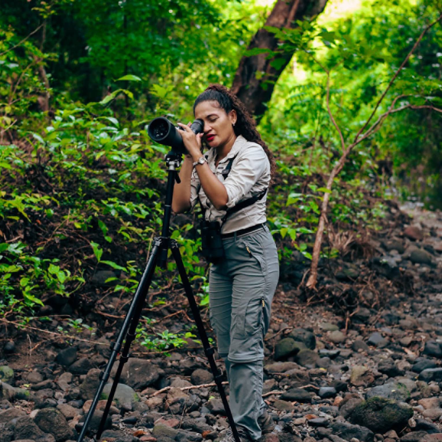 A photographer standing in a rocky area surrounded by lush foliage looks through the lens of a camera on a tripod.