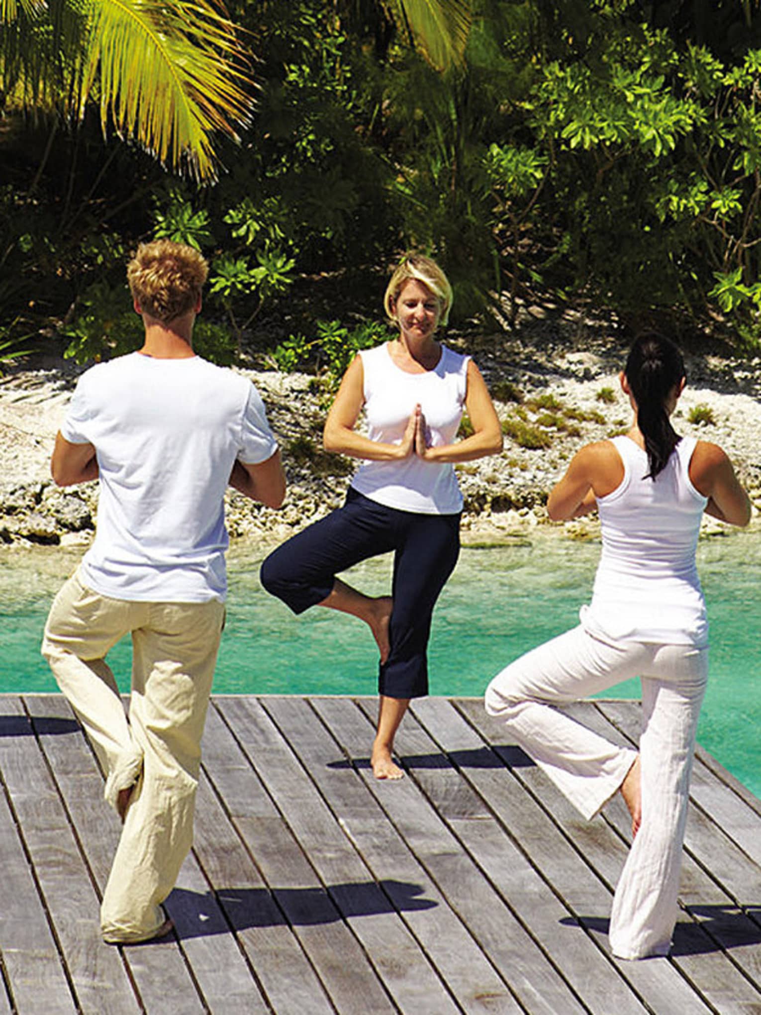 Man and two women in yoga poses on dock, standing on one leg with hands in prayer
