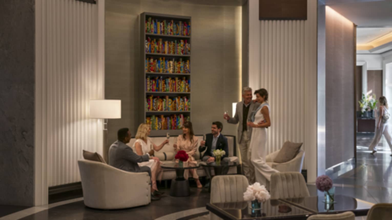 A group of people enjoy cocktails in a lobby bar in front of a large shelf lined with colourful books