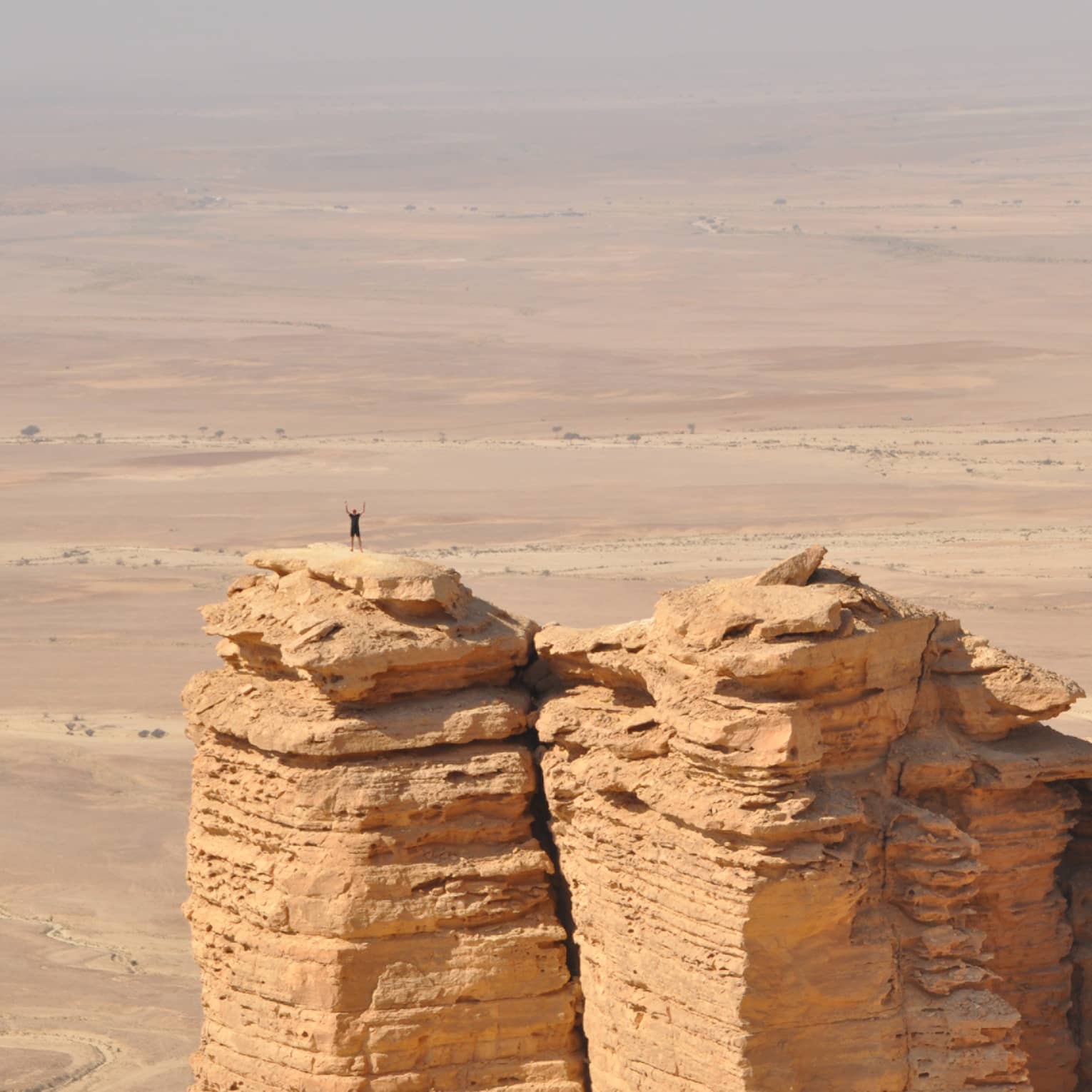An aerial view of a giant golden cliff surrounded by a dry desert and a small silhouette of a person standing on top of it.