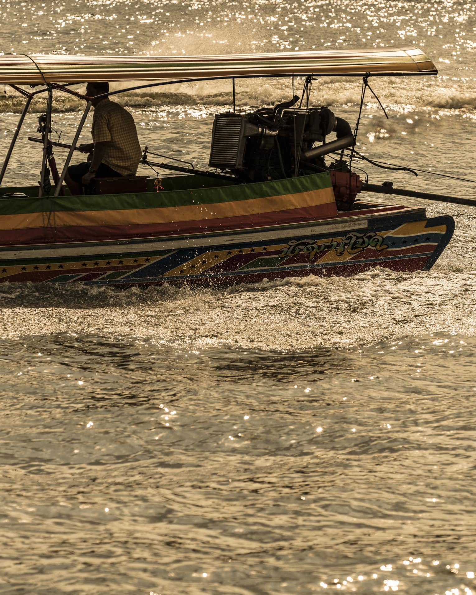 A boater sits on the edge of a colourful longtail boat, producing a frothy wake while sailing through sun-dappled water.