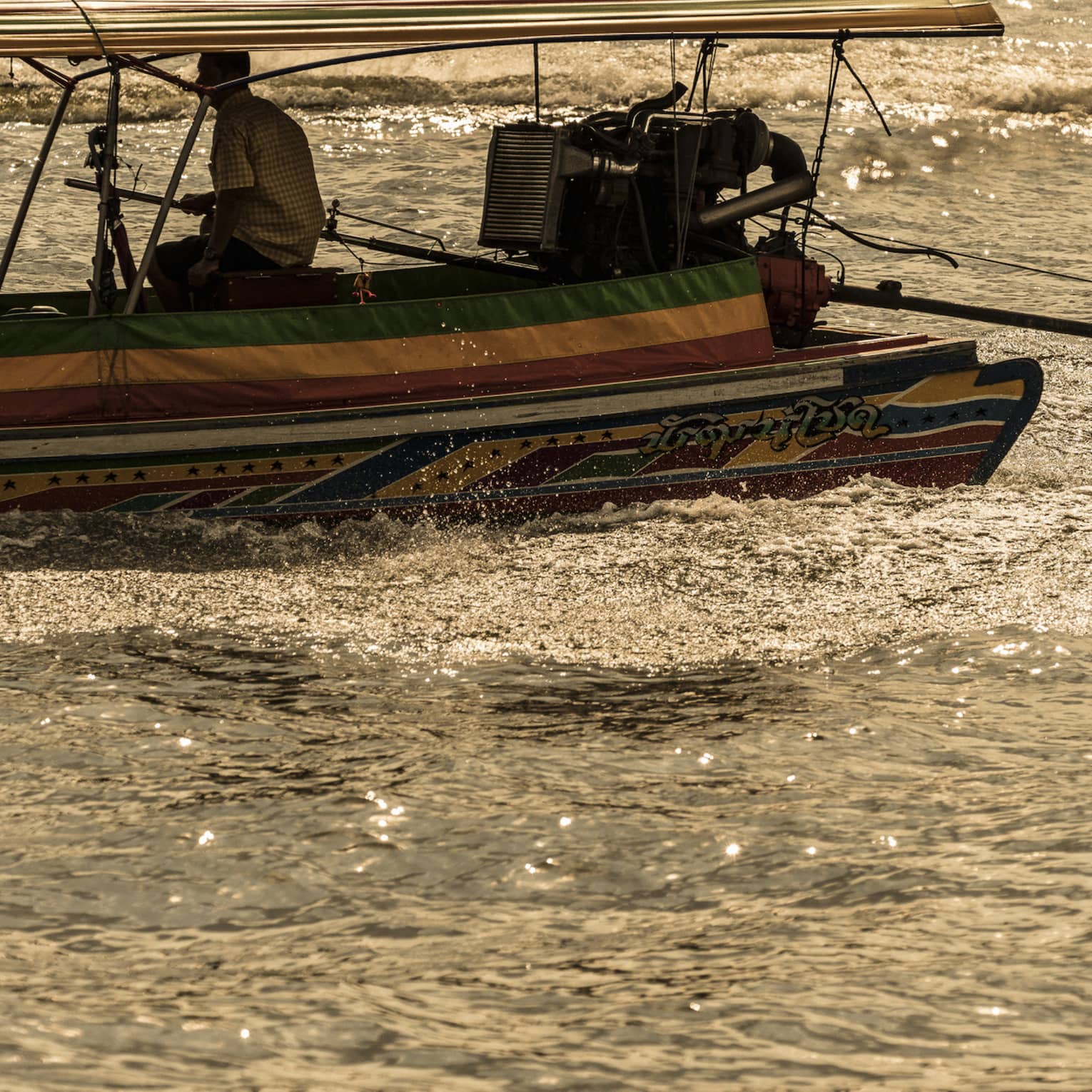 A boater sits on the edge of a colourful longtail boat, producing a frothy wake while sailing through sun-dappled water.