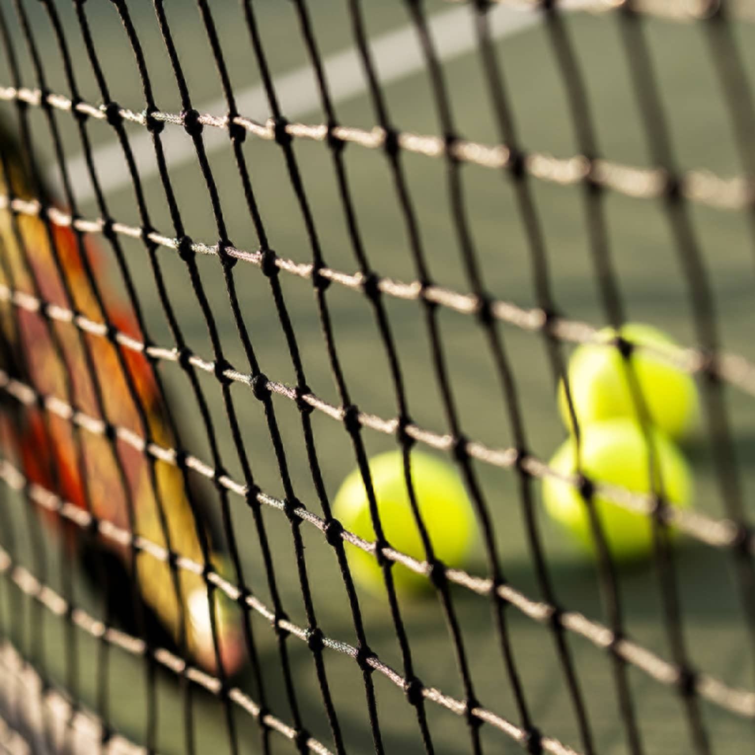 Close-up of tennis racket against net, three green tennis balls on ground