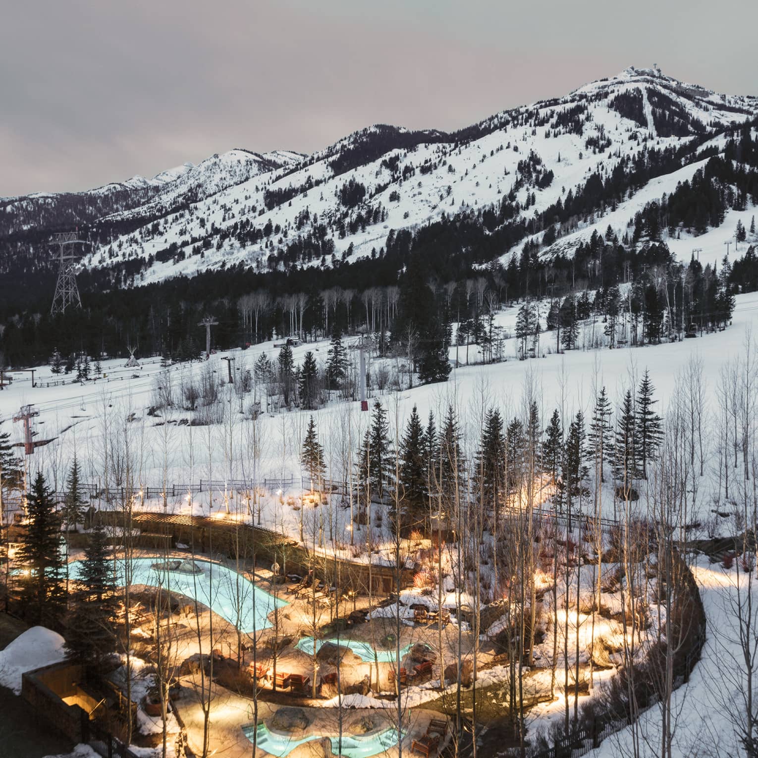 Aerial view of illuminated blue outdoor swimming pool through trees on snowy hill at dusk