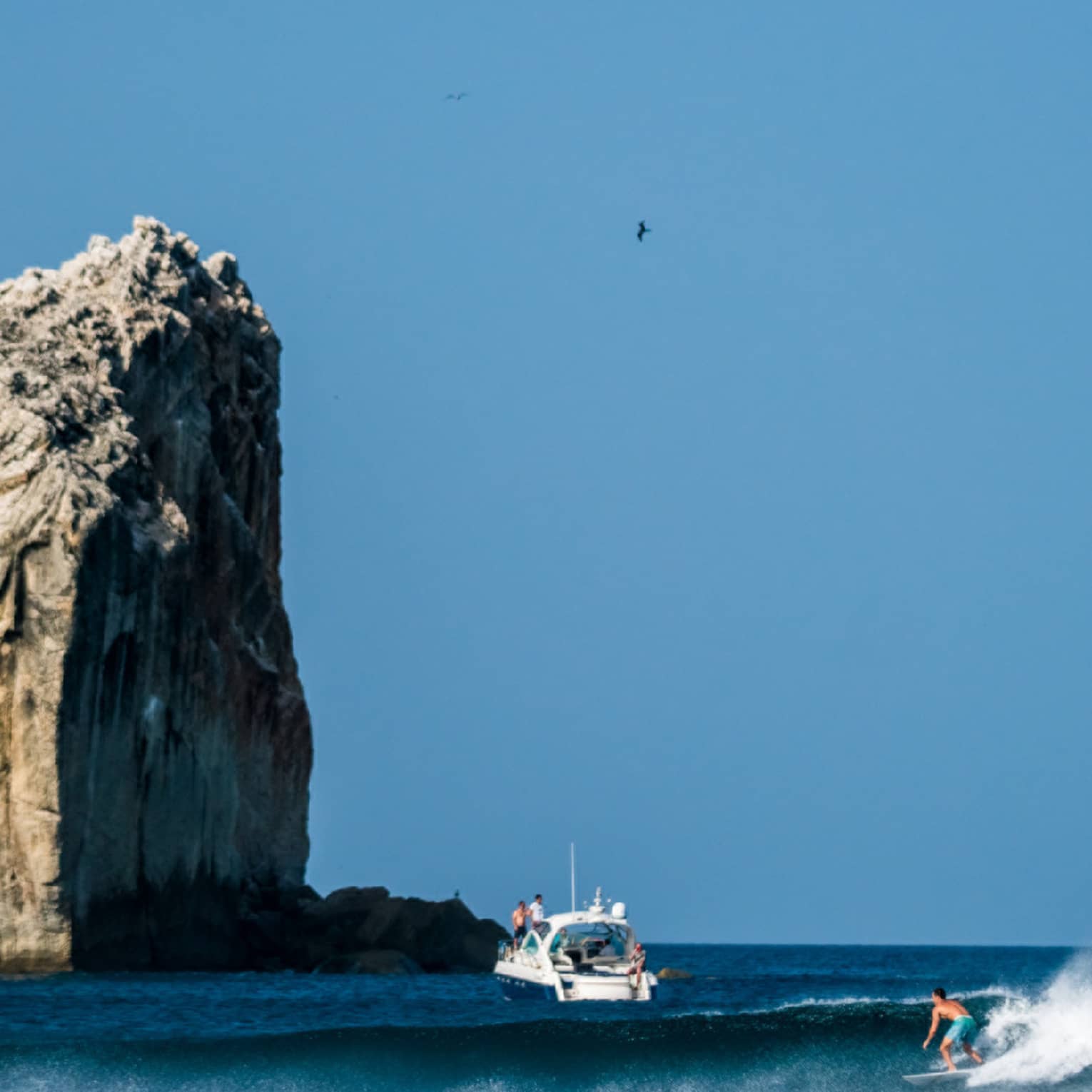 A surfer rides a wave in the foreground, with a boat nearby and a large rock formation towering above the water in the background