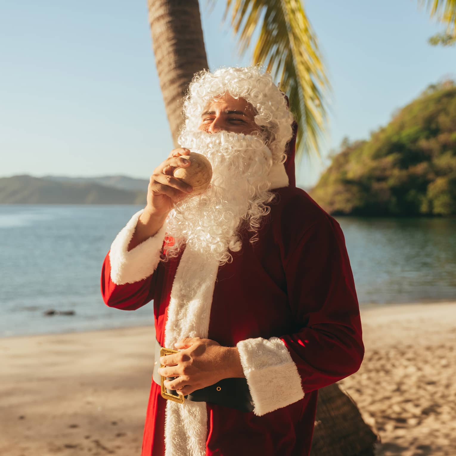A person on the beach wearing a Santa Claus costume and holding a coconut