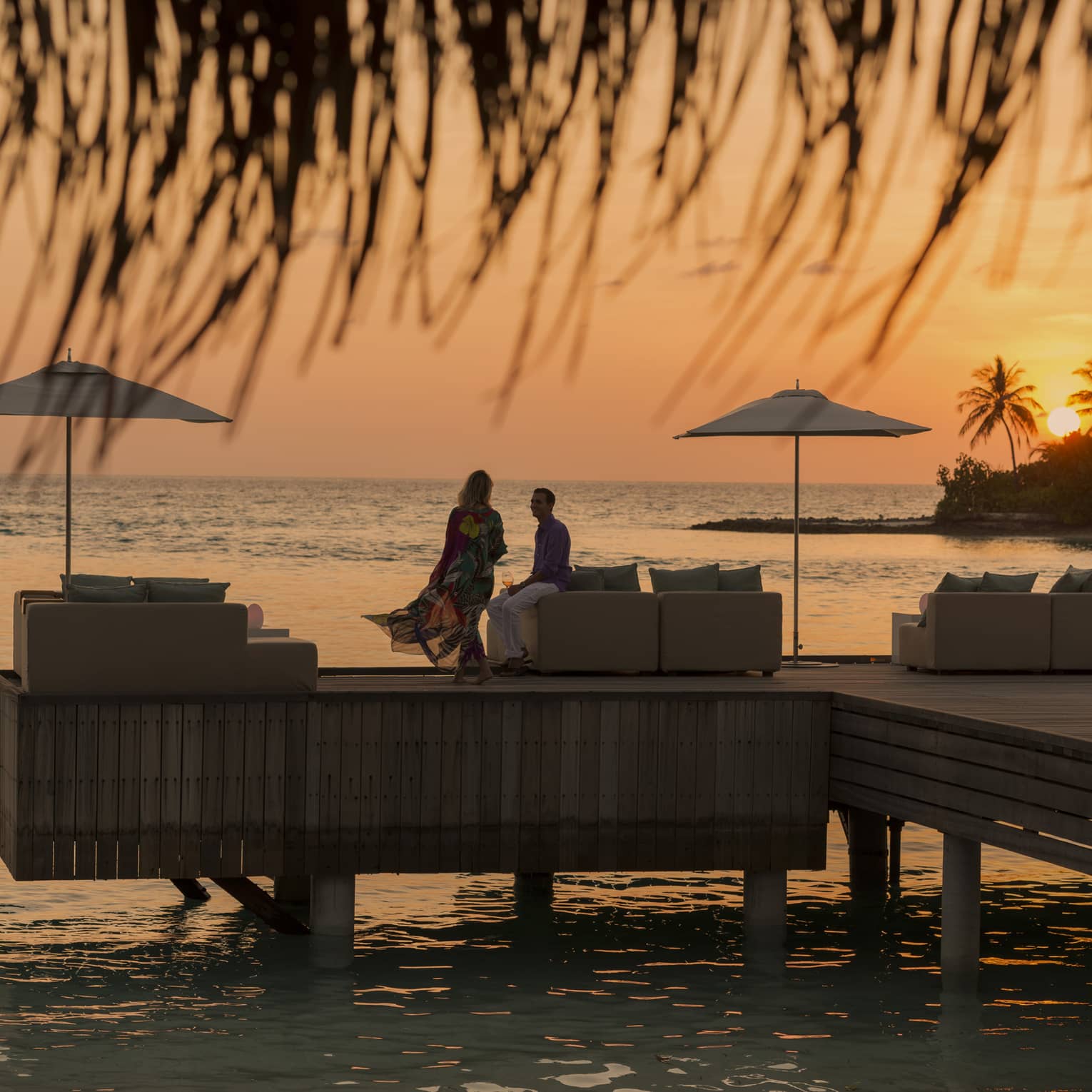 Private island dining at sunset. Woman with long dress blowing in the wind, man sits on the edge of white sofa