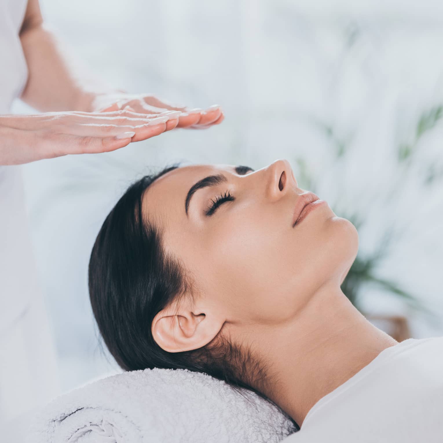 Woman with eyes closed lays on rolled up towel on spa table under therapists' hovering hands