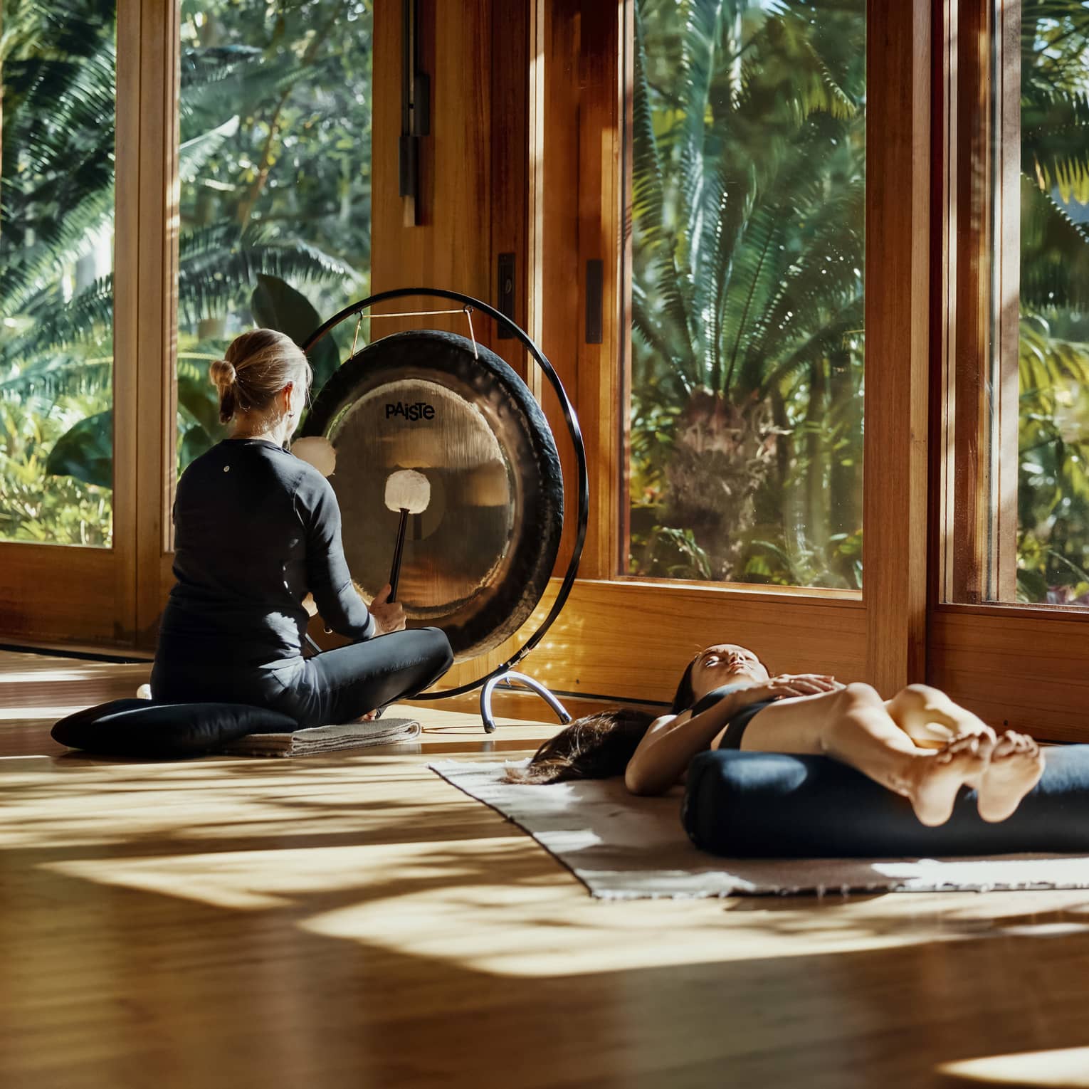 During a sound bath, a meditator lays on a mat, legs resting on a bolster, as a practitioner plays a gong with two mallets.