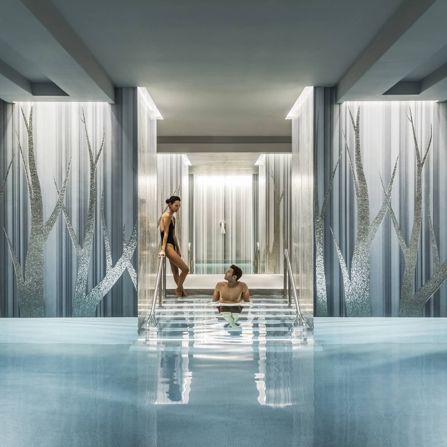 Woman in black swimsuit enters serenity pool while man sits on steps between blue and silver columns
