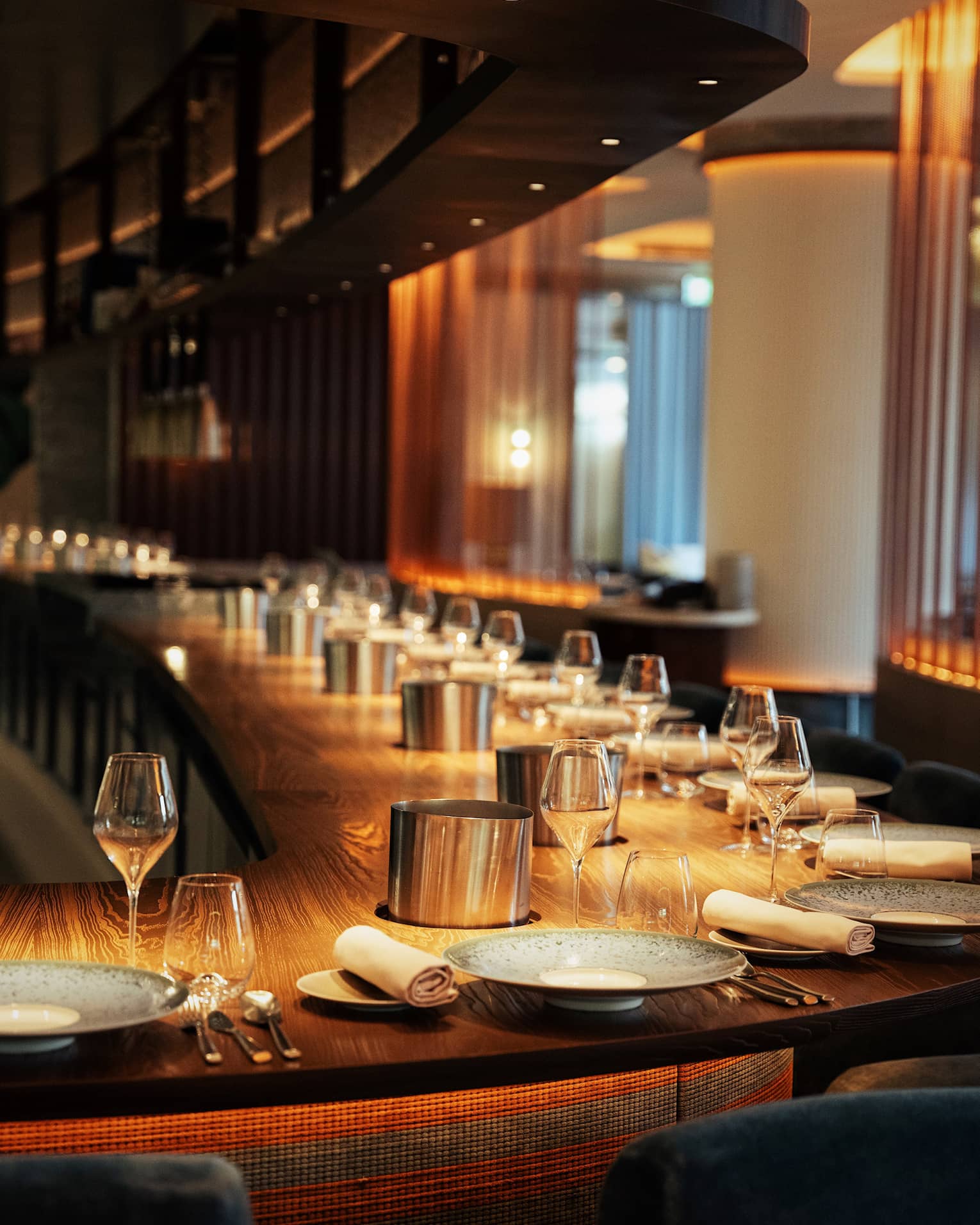 A long, curved solid wood bar with a dozen place settings in warm, golden lighting.